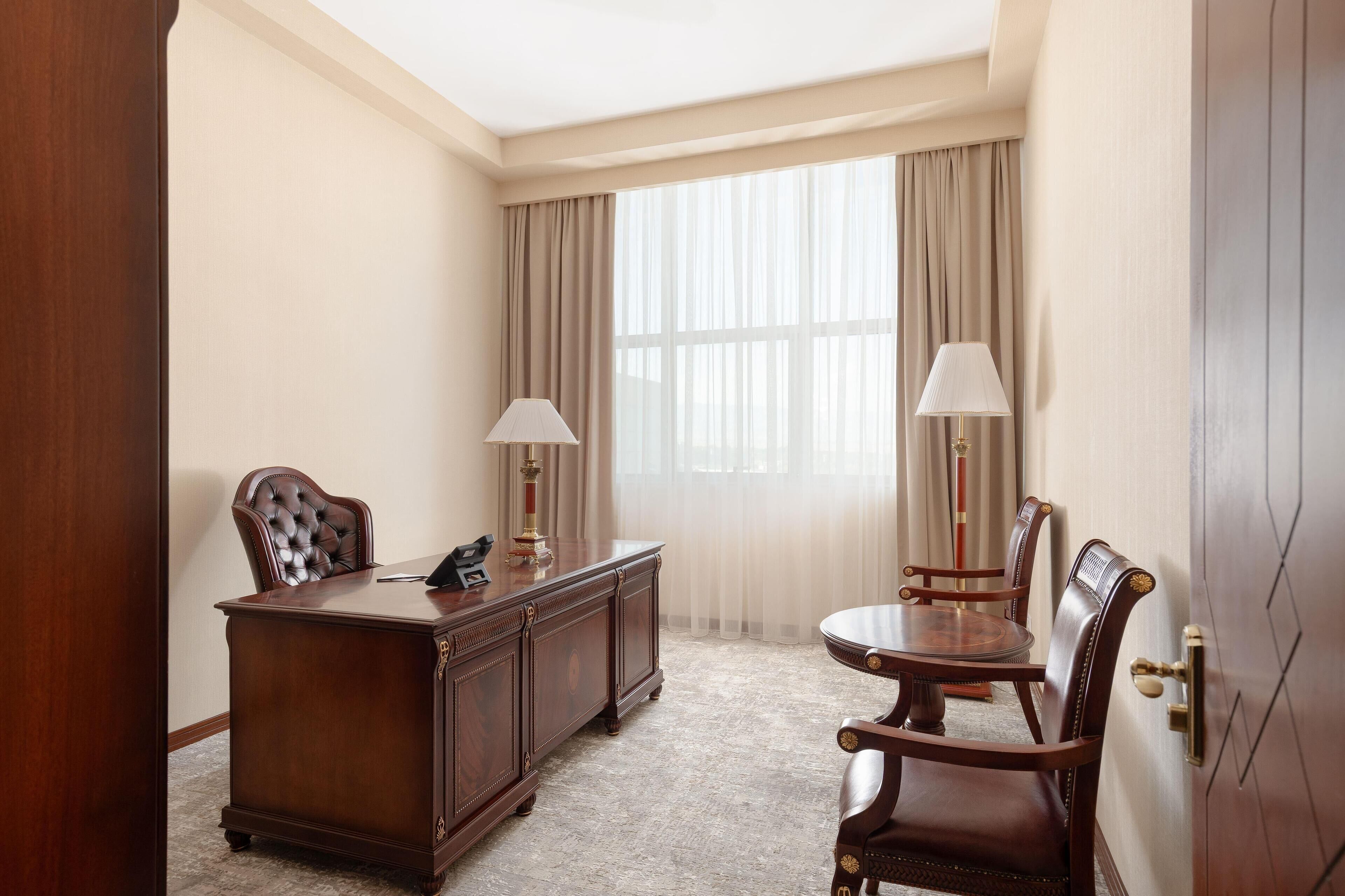 A room with a wooden desk, two chairs, a lamp, and a large window with curtains.