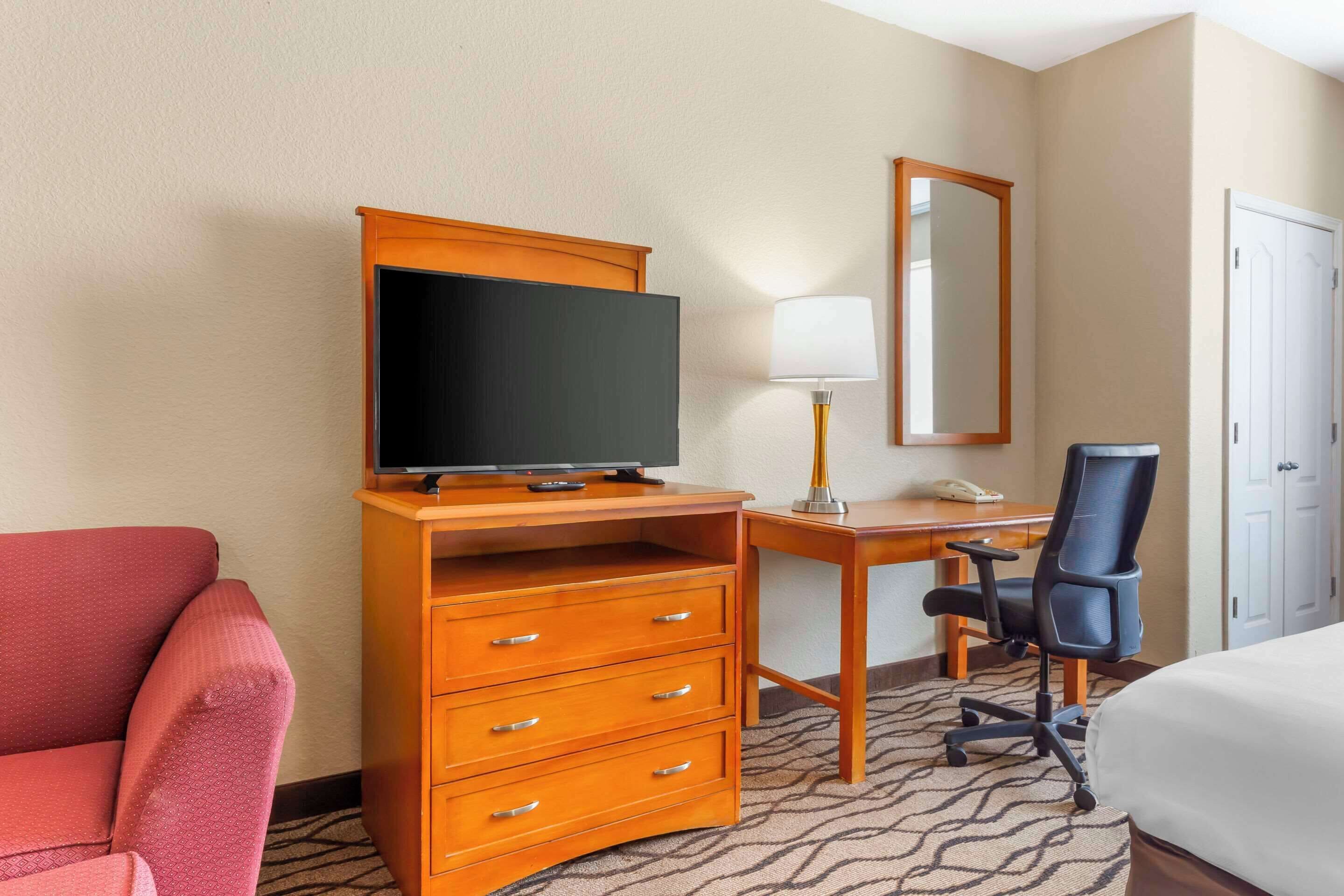 A hotel room with a wooden desk, a television, a chair, a bed, and a red armchair.