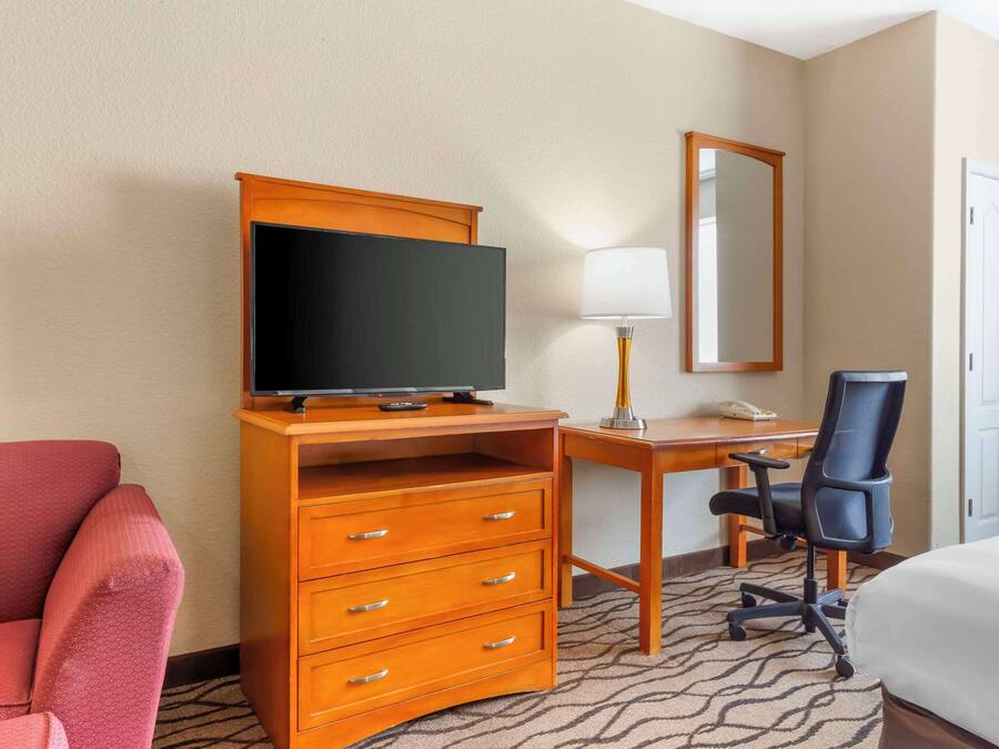 A hotel room with a wooden desk, a television, a chair, a bed, and a red armchair.