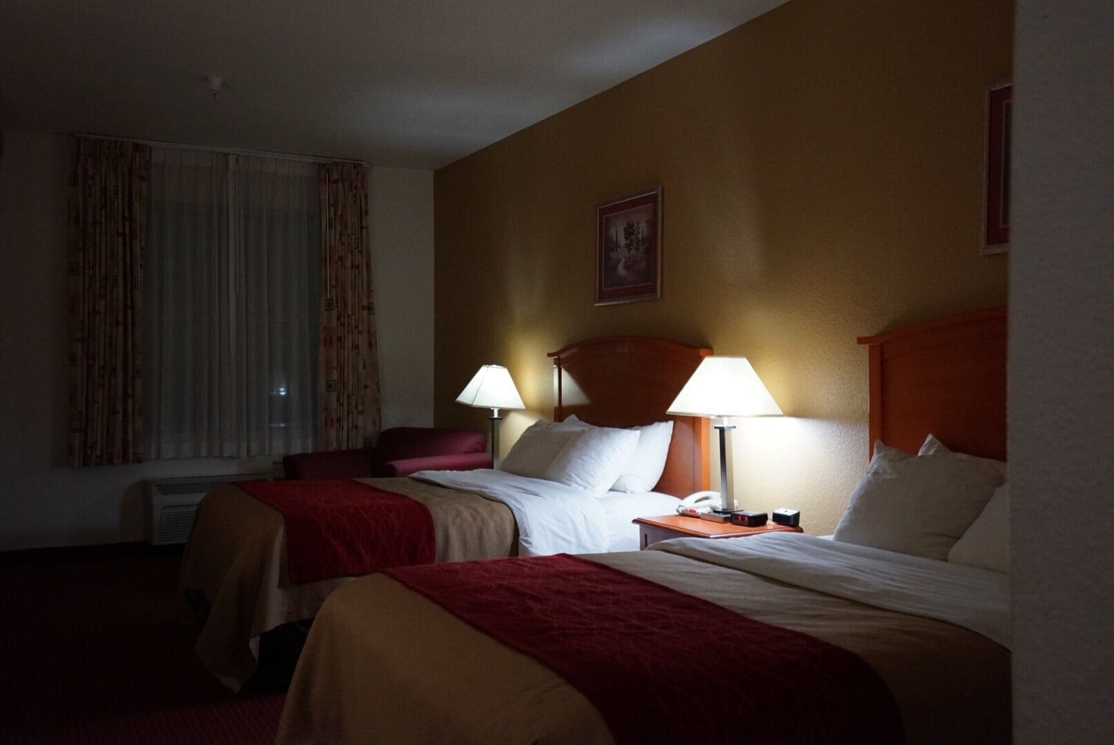 A hotel room with two beds, a red chair, a window with curtains, and a wall-mounted lamp.