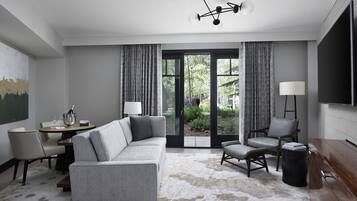 En moderne stue med en grå hjørnesofa, et lille rundt bord med en flaske og glas, en stol, en lampe og et fjernsyn på væggen.