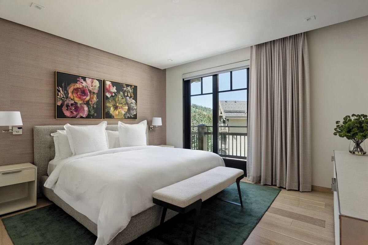 Ein modernes Hotelzimmer mit einem großen Bett, einer Bank, zwei Bildern, einem Nachttisch und Blick ins Freie.