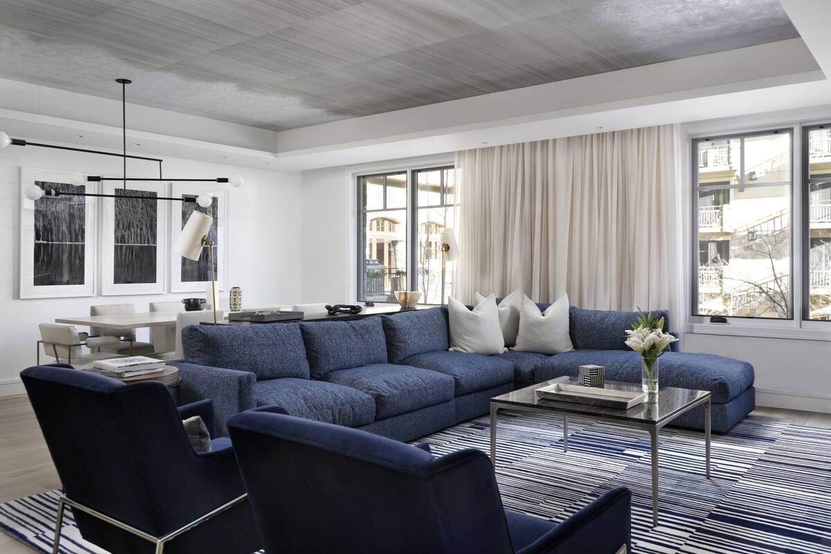 Ein modernes Wohnzimmer mit einem blauen modularen Sofa, einem Couchtisch und einem Streifenmuster-Teppich. Zwei Sessel, ein Esstisch und eine Deckenleuchte sind ebenfalls vorhanden.