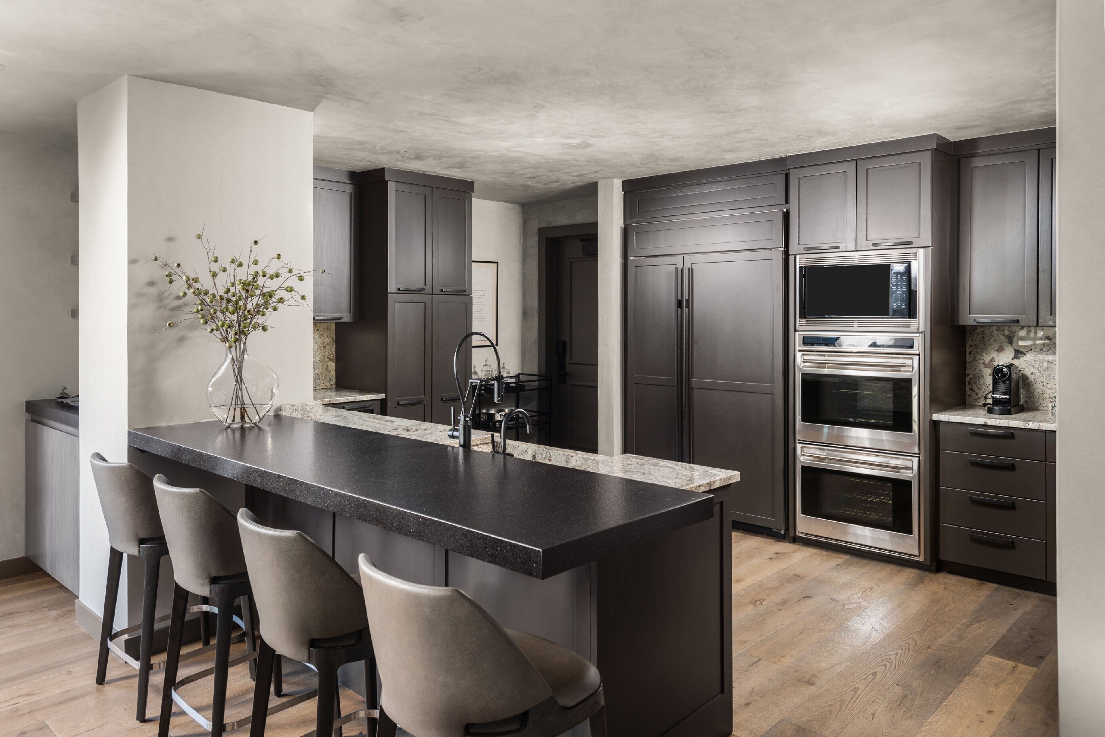 A modern kitchen with a central island, bar stools, dark cabinetry, stainless steel appliances, and a vase with branches.