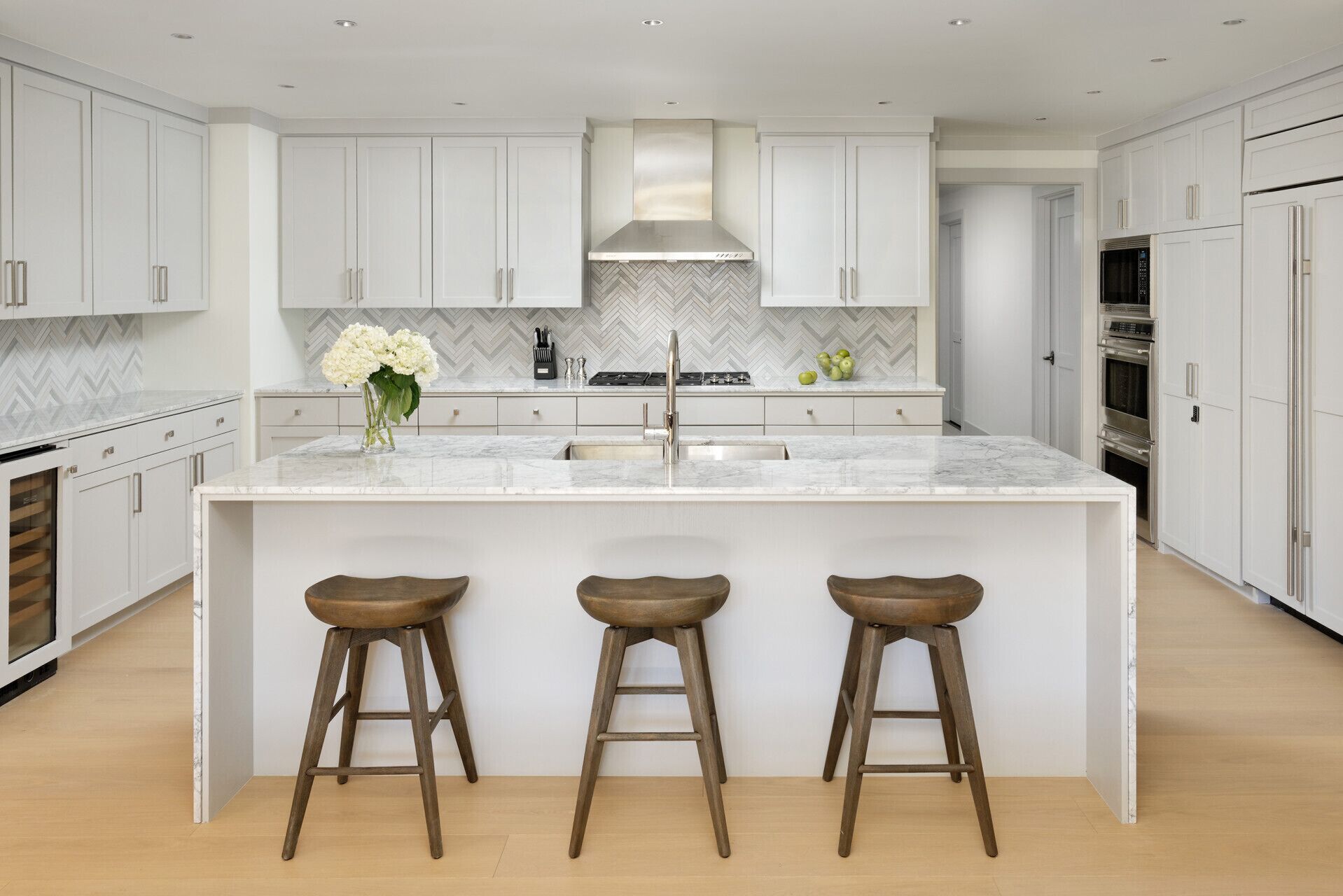 A modern kitchen with white cabinetry, a marble countertop, a central island with stools, and stainless steel appliances.