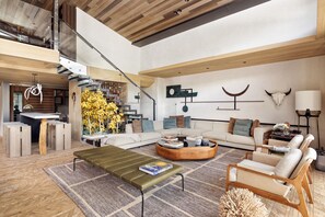 A spacious living area with a modern design, featuring a wooden coffee table, a sofa, and a unique curved seating area. A staircase leads to an upper level, and there are decorative elements like a skull and a crescent moon on the wall.