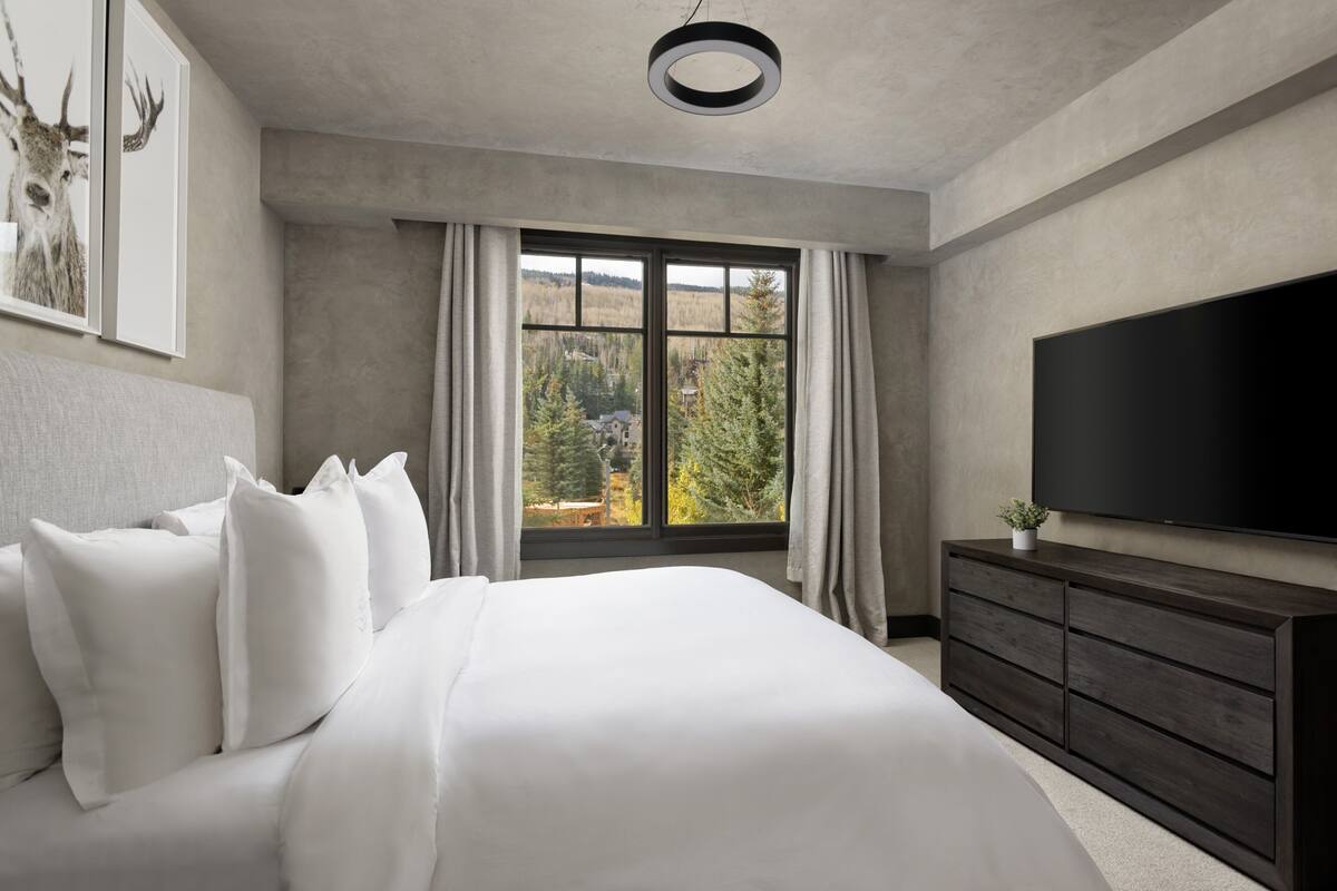 Ein modernes Schlafzimmer mit einem großen Bett, einer Kommode, einem Flachbildfernseher und einem Fenster mit Blick auf den Wald.
