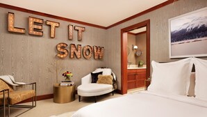 A modern hotel room with a large bed, two armchairs, a round side table, and a wall-mounted sign that reads "LET IT SNOW".