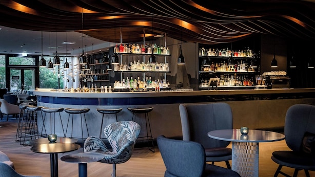 A bar area with multiple seating options, a bar counter with shelves of bottles, and a modern ceiling design. - Motel One Hamburg-Alster (Hamburg)