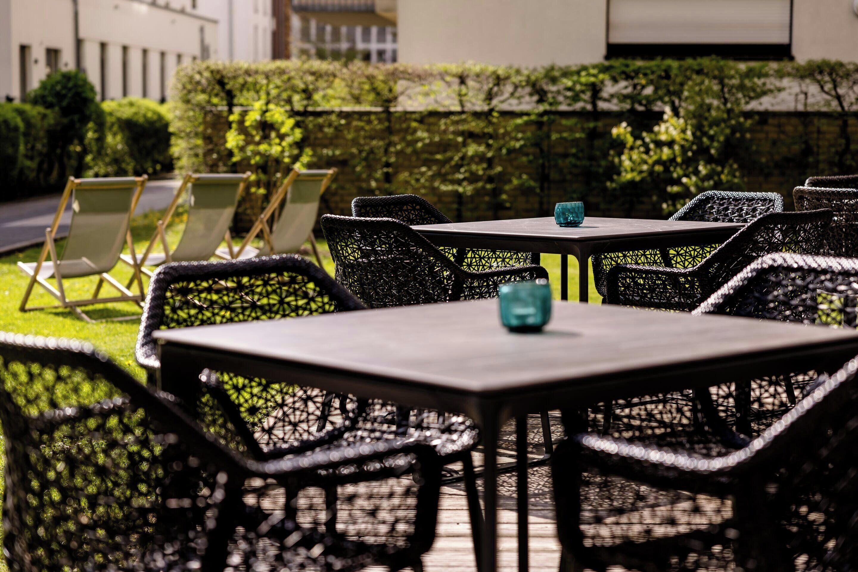 An outdoor seating area with wicker chairs and tables, a hedge, and a building in the background.