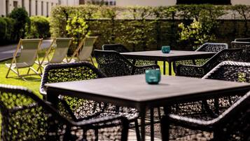 An outdoor seating area with wicker chairs and tables, a hedge, and a building in the background.