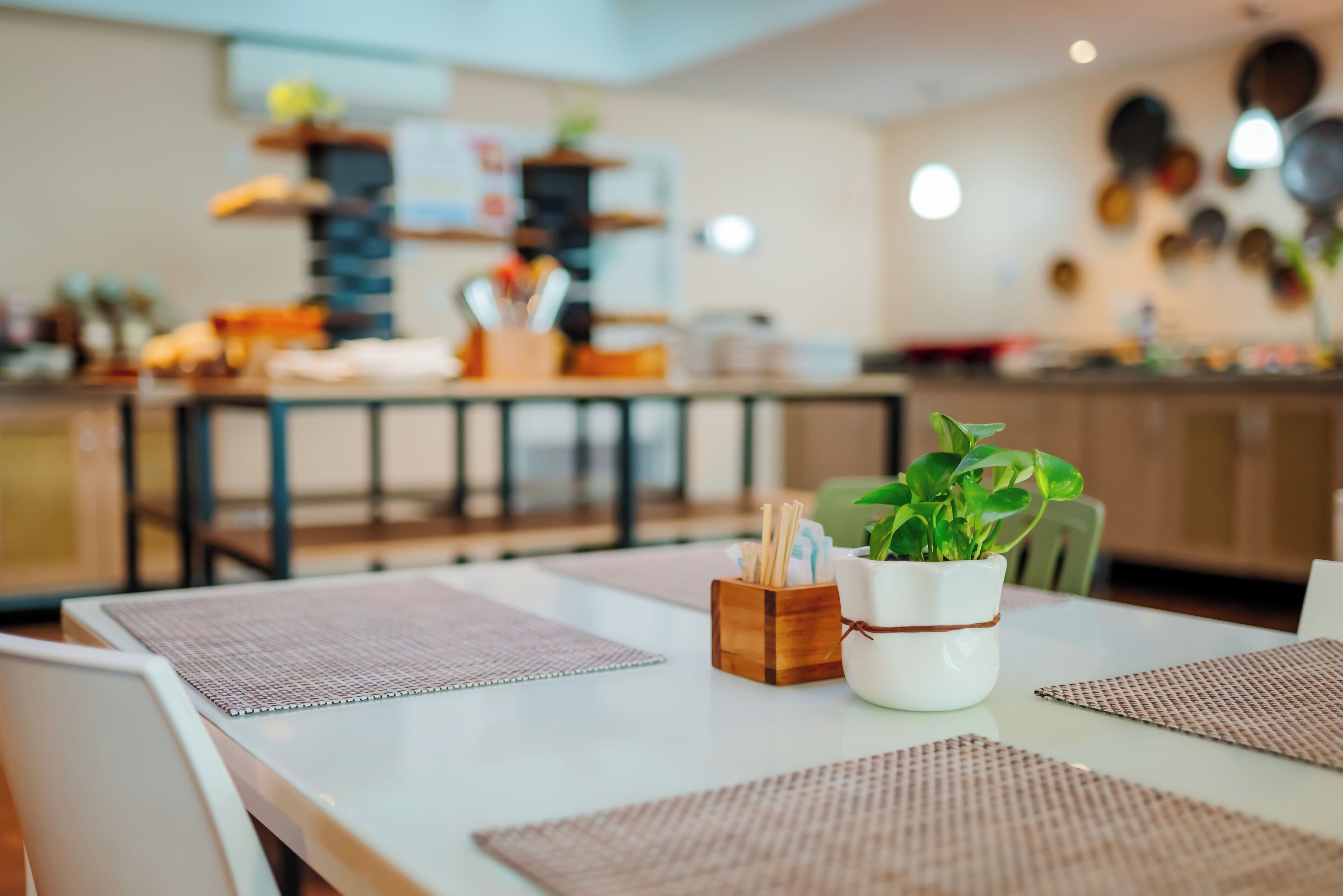 A dining area with a white table, brown placemats, a white vase with a green plant, and a wooden utensil holder.