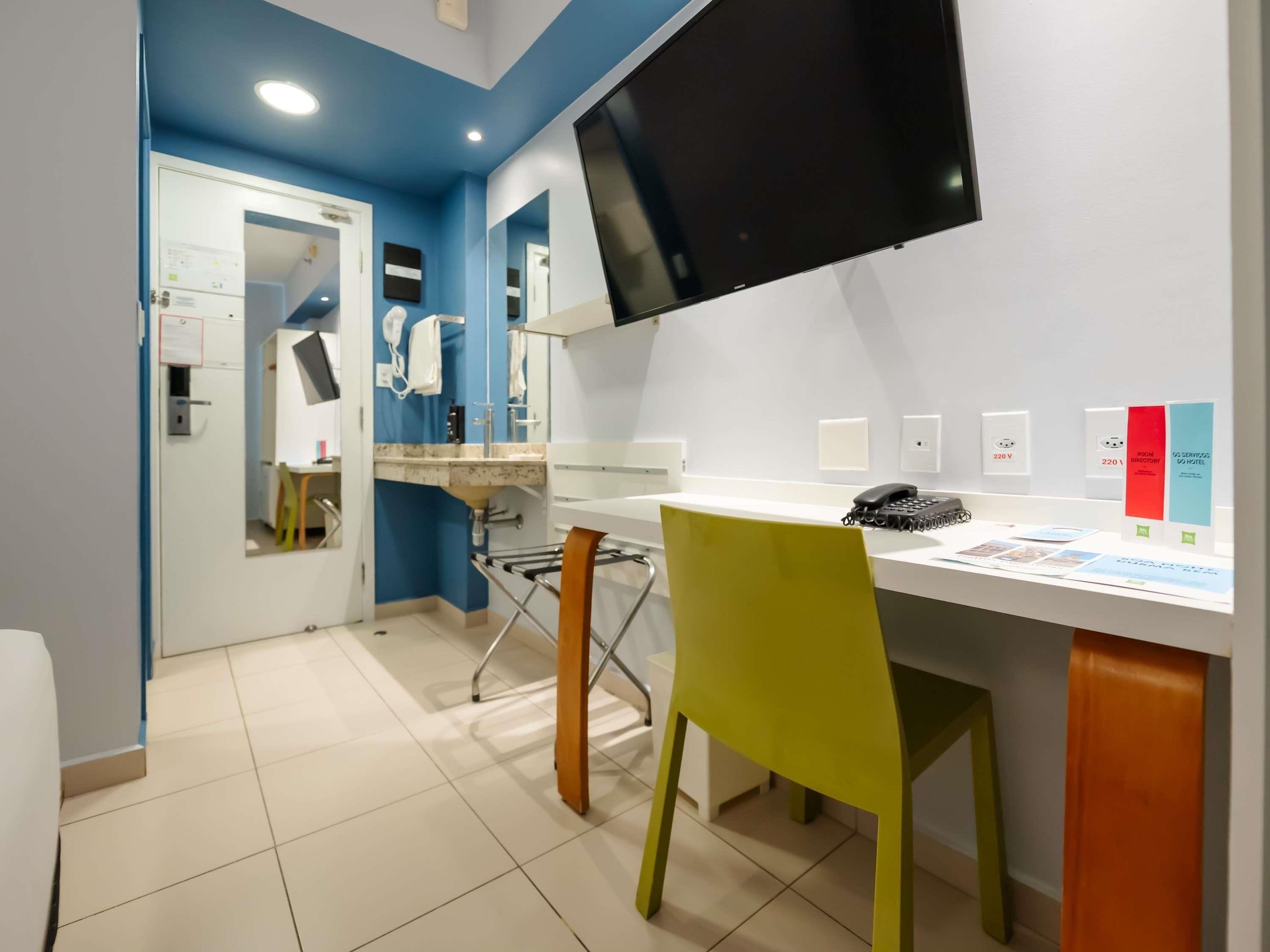 A hotel room with a desk, chair, flat-screen TV, and a mirror.