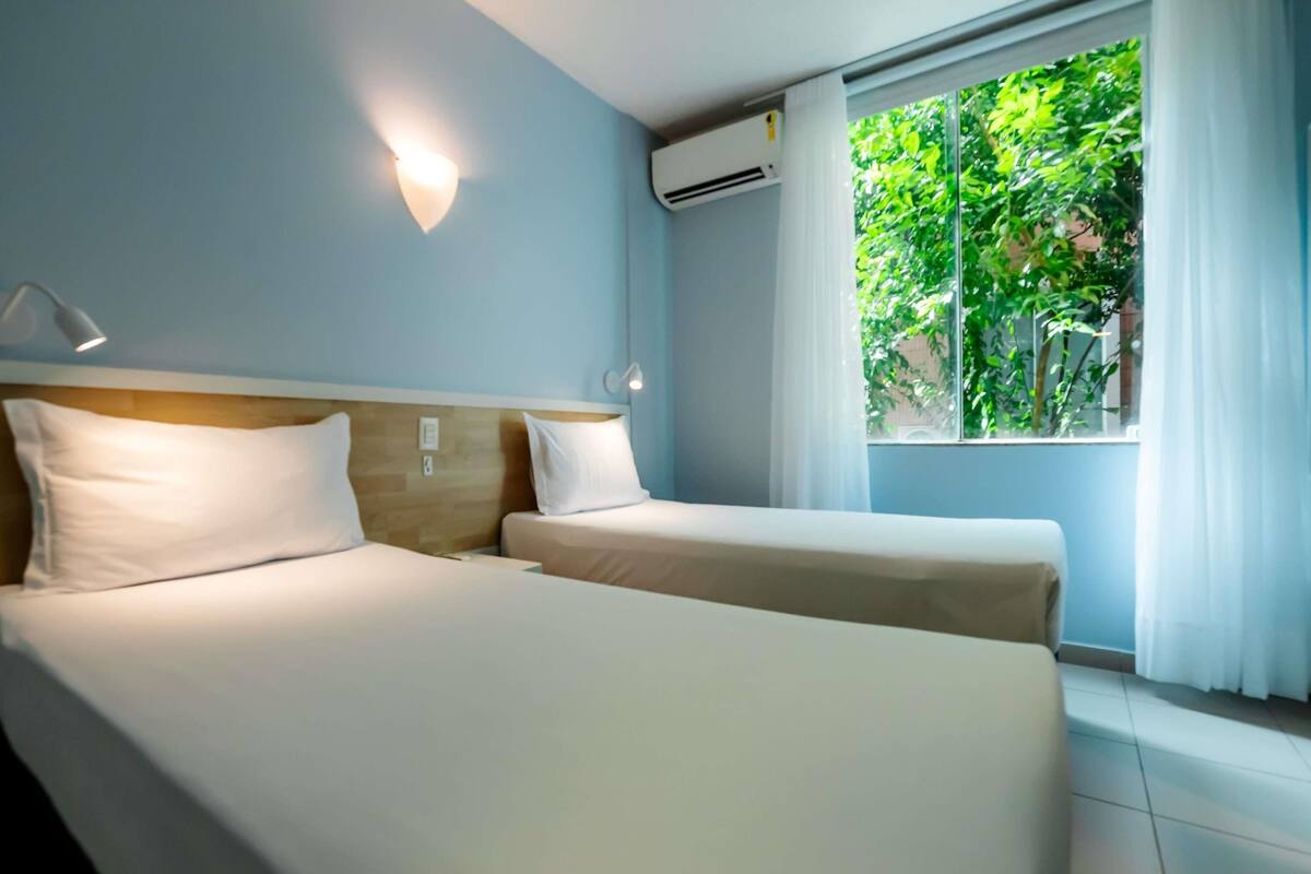 A hotel room with two beds, a window with a view of greenery, an air conditioning unit, and a wall-mounted light fixture.