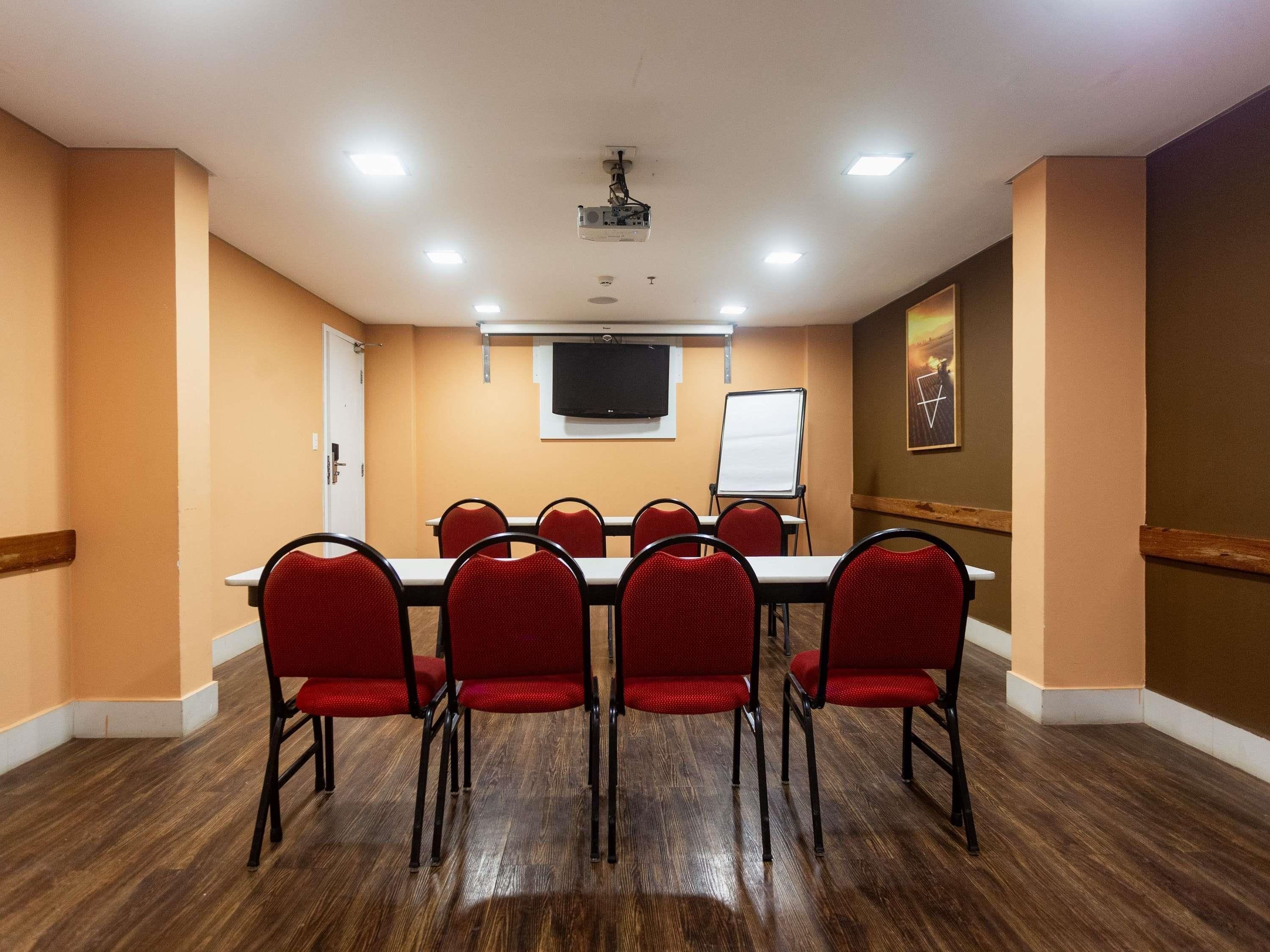 A conference room with a long table, red chairs, a whiteboard, a projector, and a TV on the wall.
