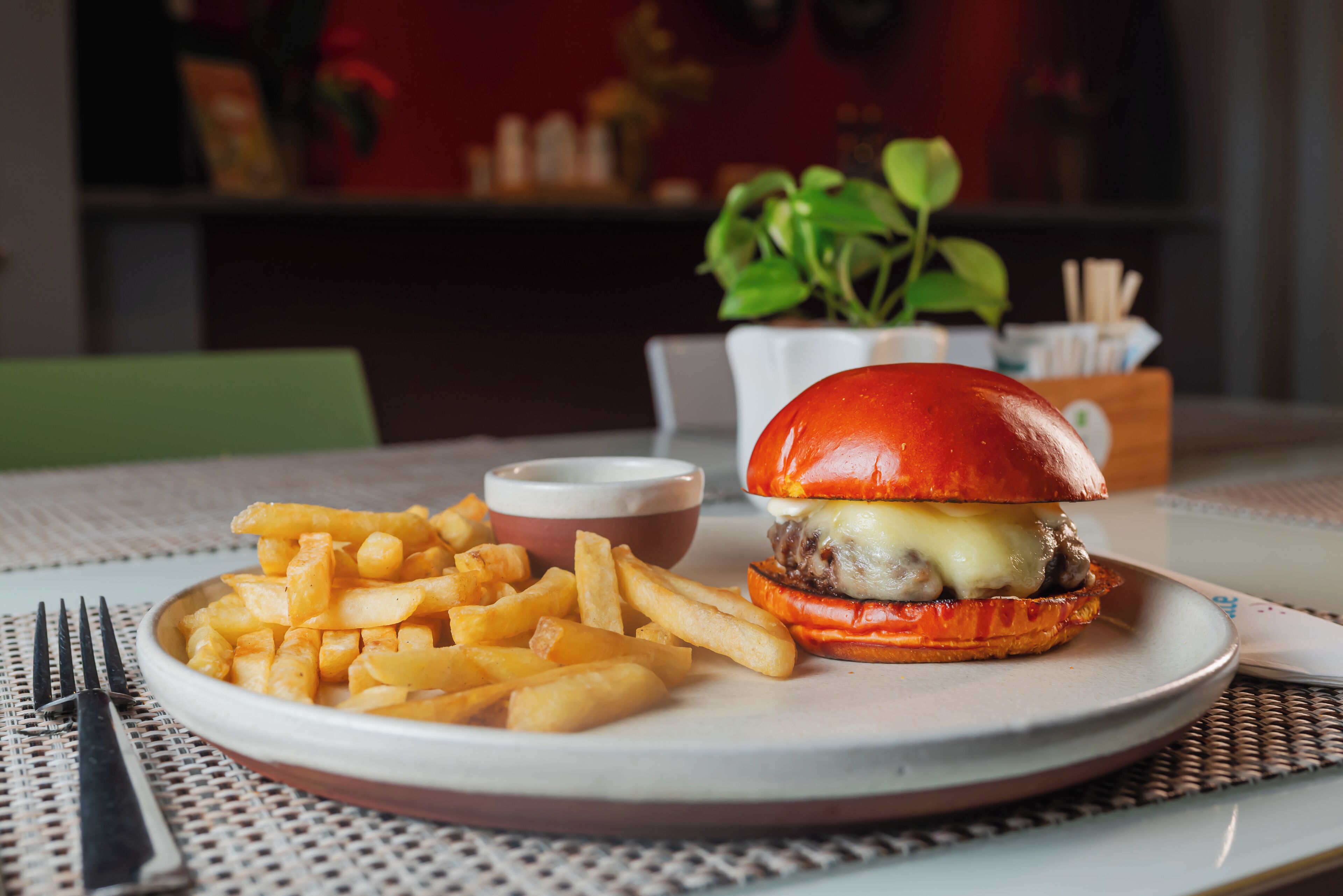 Un plato con una hamburguesa y papas fritas, un cuenco pequeño de salsa y un tenedor sobre una mesa.