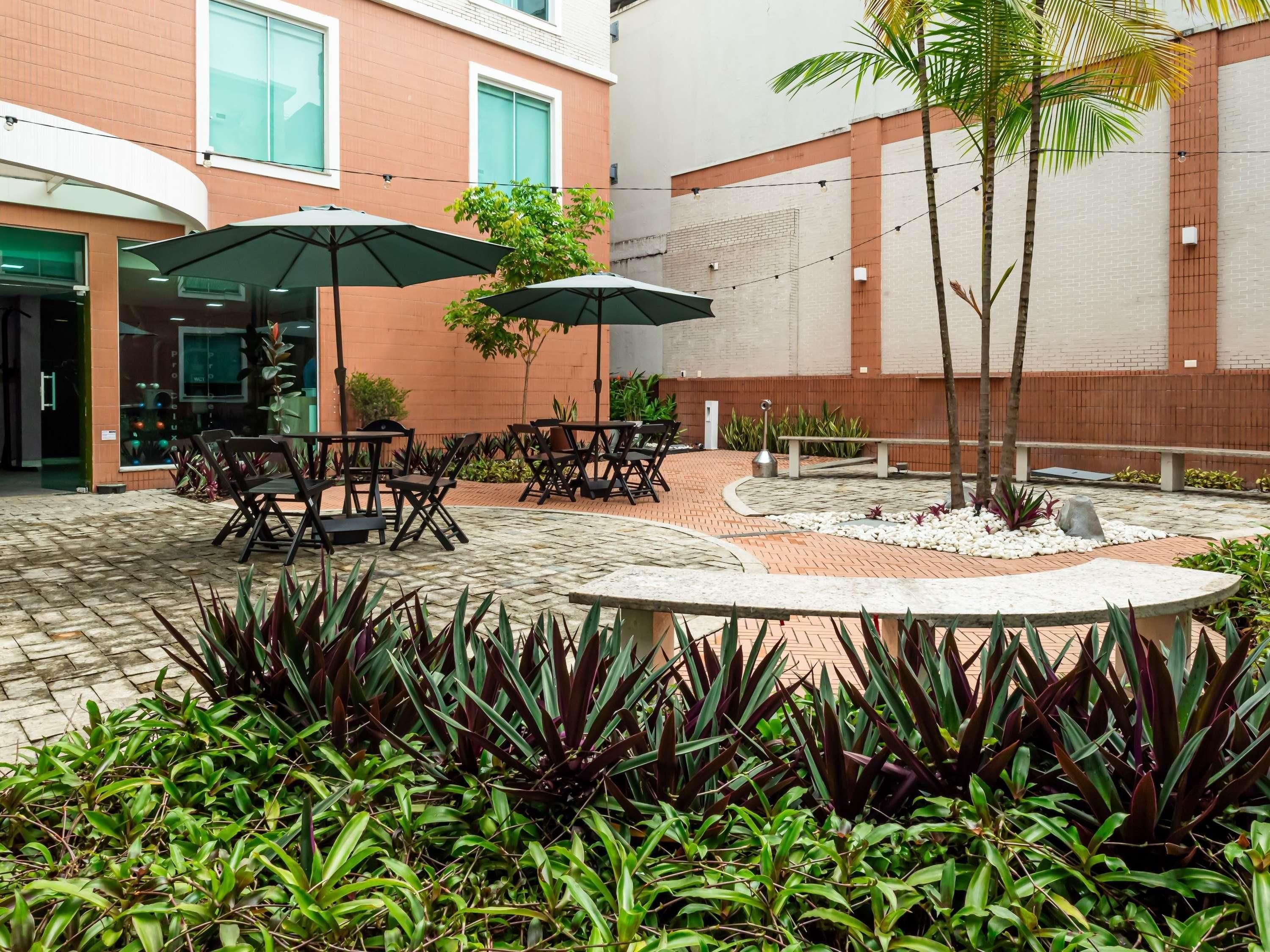 Un patio al aire libre con mesas y sillas, rodeado de plantas y un elemento decorativo circular de piedra.