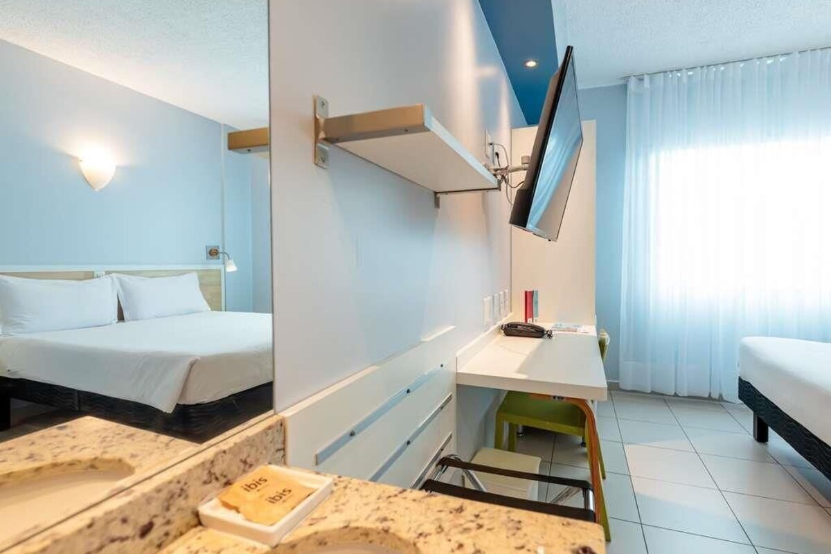 A hotel room with two beds, a desk, a television, and a bathroom with a sink and mirror.