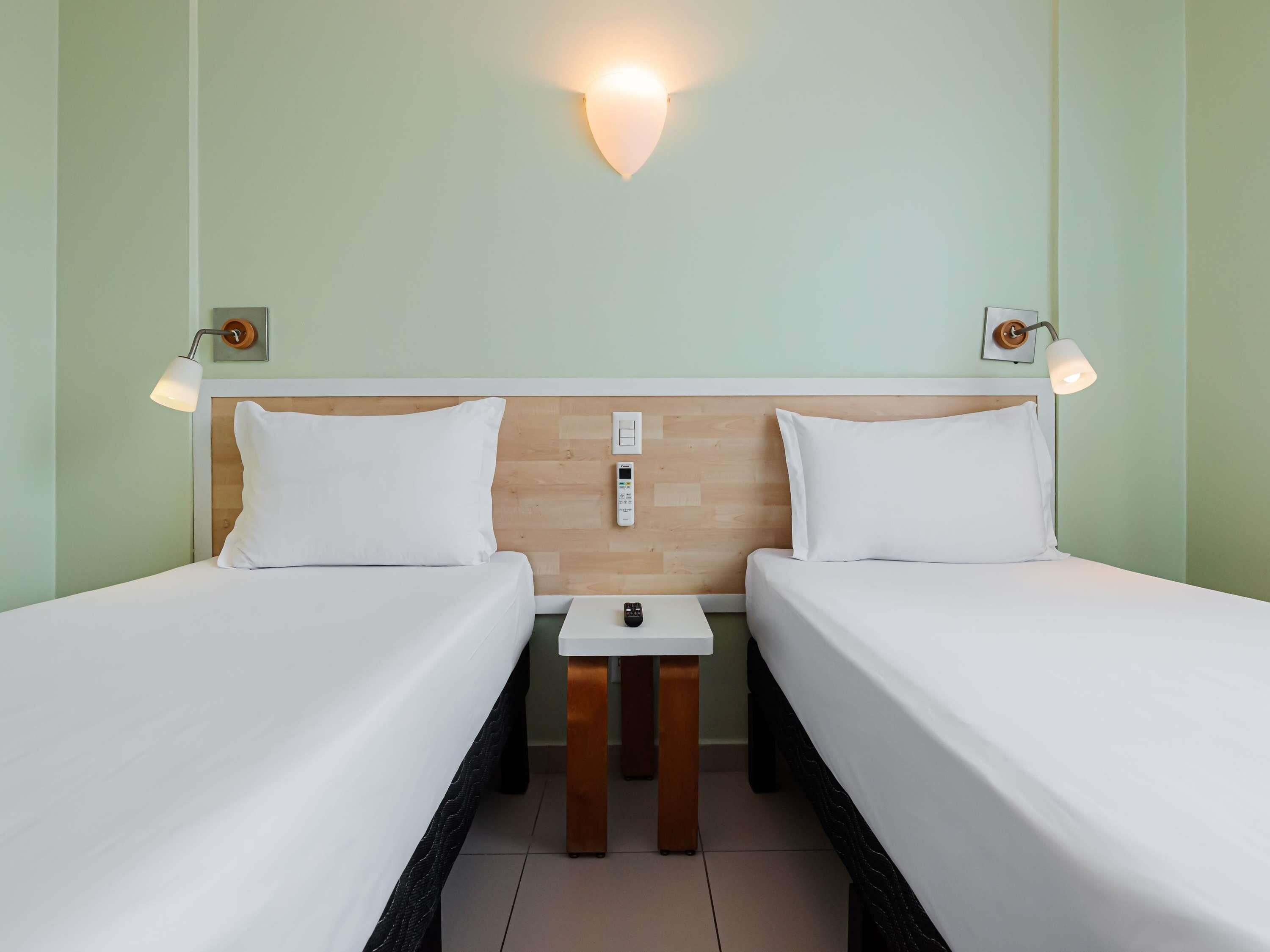 A hotel room with two single beds, a wooden headboard, bedside tables, and wall-mounted lamps.