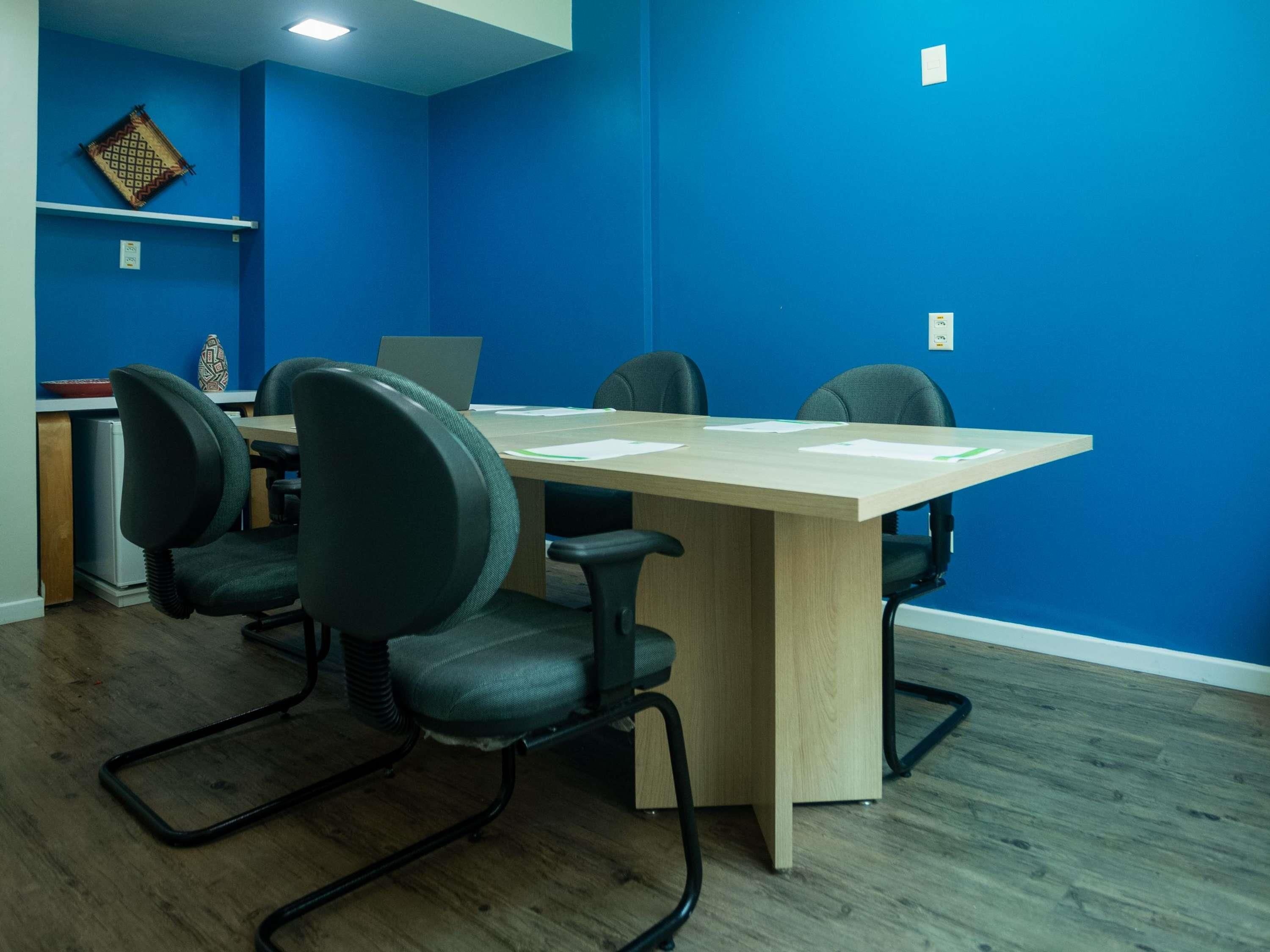 Una sala de conferencias con una mesa larga, cuatro sillas, una computadora portátil y un jarrón decorativo en un estante.
