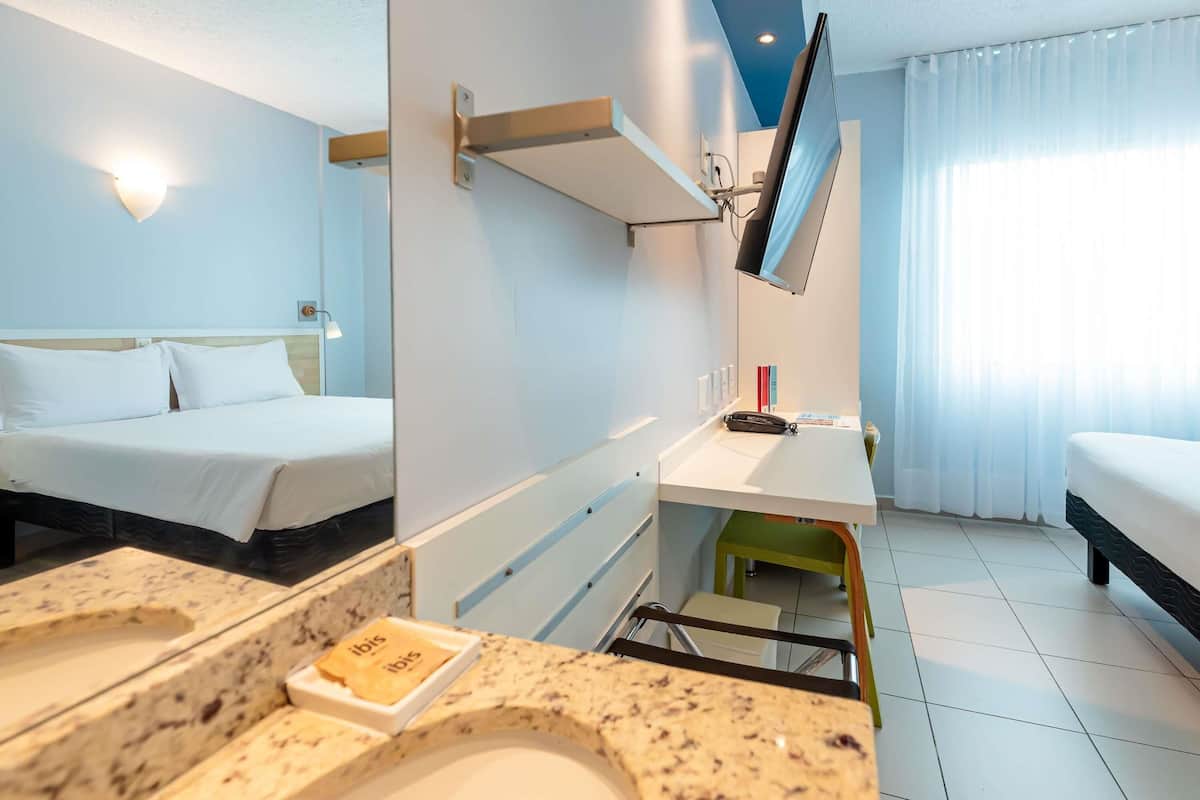 A hotel room with two beds, a desk, a mirror, a TV, and a bathroom with a sink and toiletries.