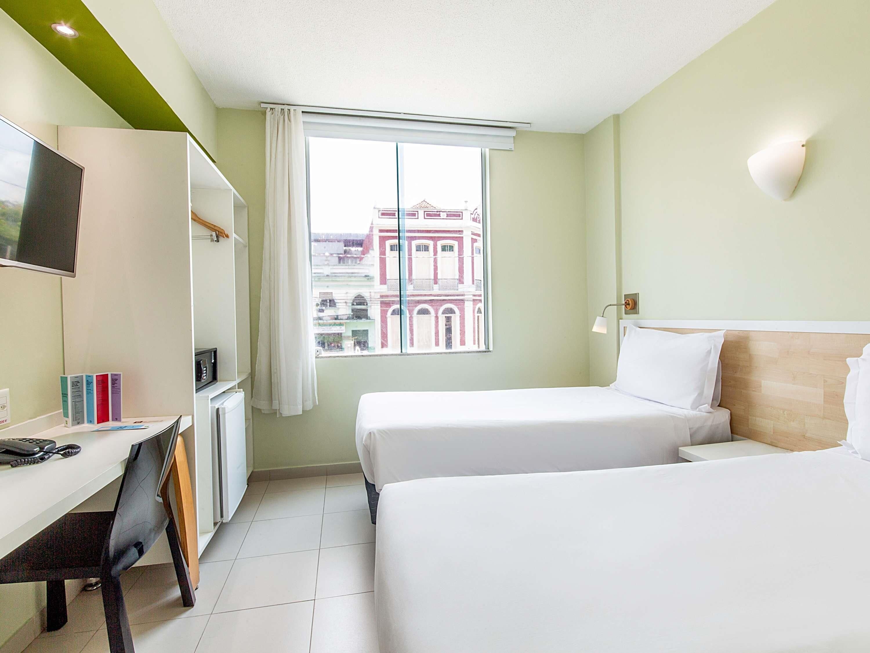 Ein Hotelzimmer mit Bett, Schreibtisch, Stuhl, Fernseher und einem Fenster mit Blick auf Gebäude.