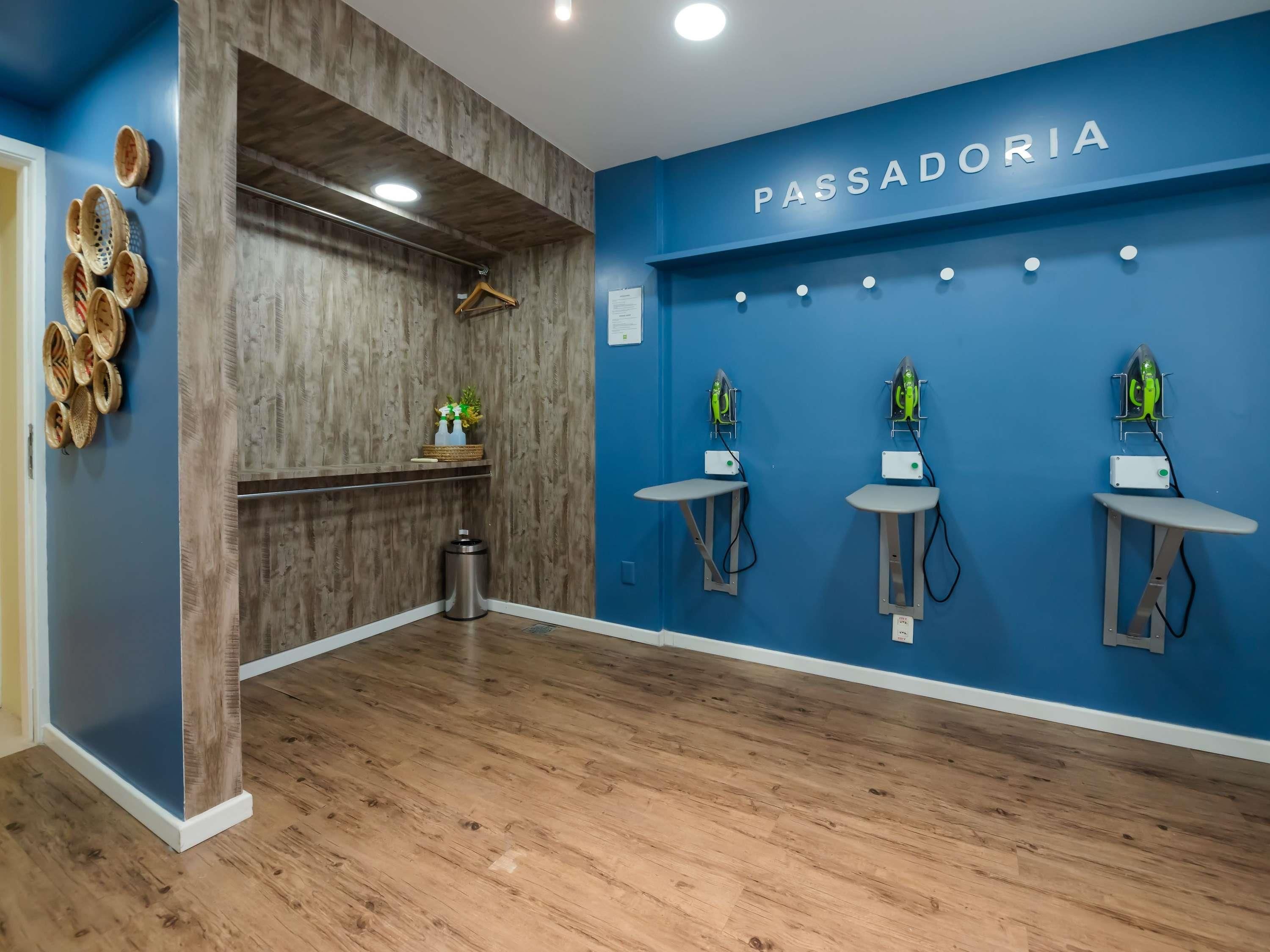 Une pièce avec un mur bleu, un plancher en bois et une enseigne sur laquelle on peut lire « PASSADORIA ». On y trouve trois postes de repassage équipés de fers à repasser et une poubelle.