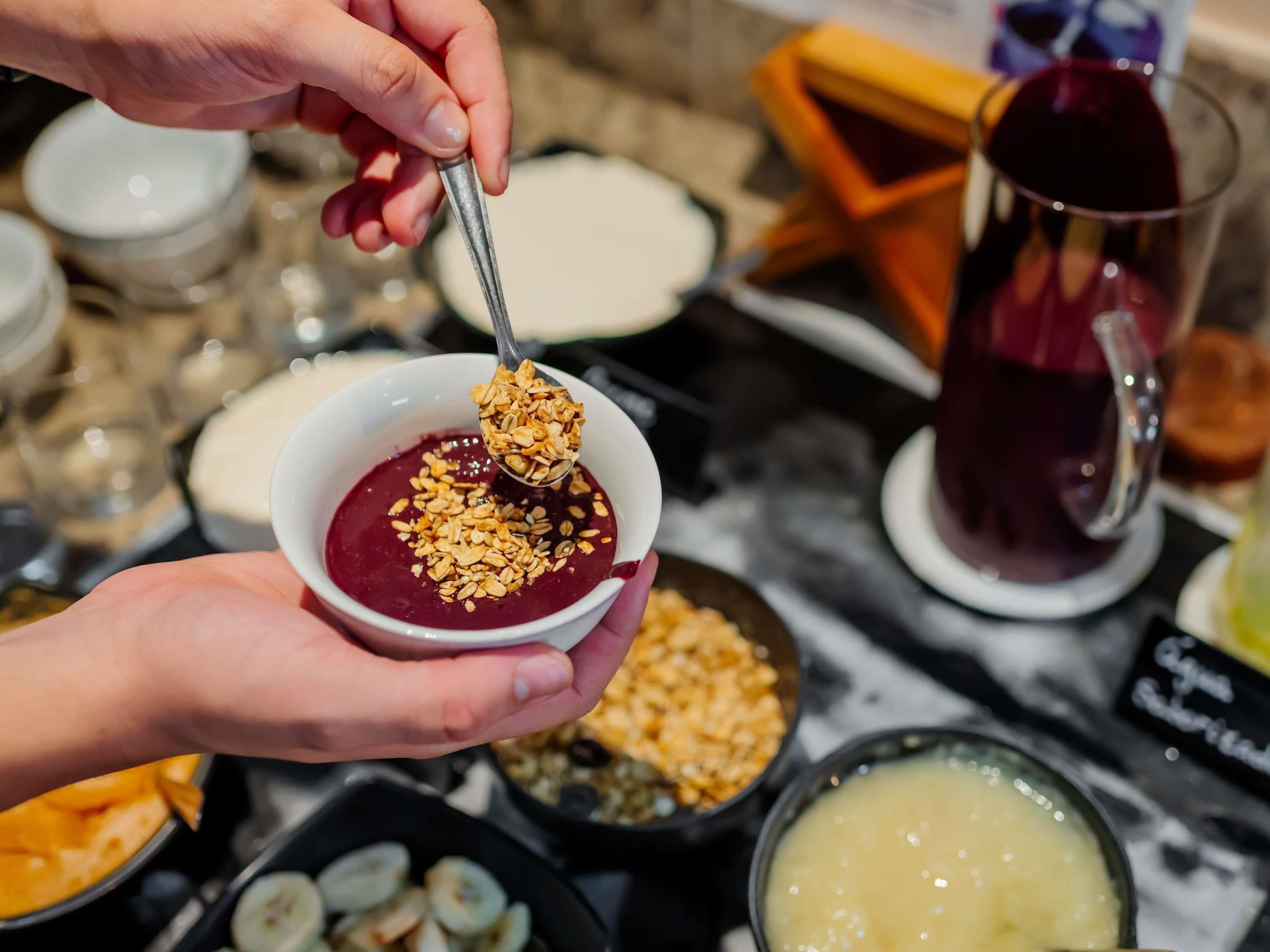Une personne tient un bol contenant une cuillère de granola au-dessus d’une substance rouge, qui pourrait être un dessert, et on aperçoit divers autres plats ainsi qu’une carafe à l’arrière-plan.