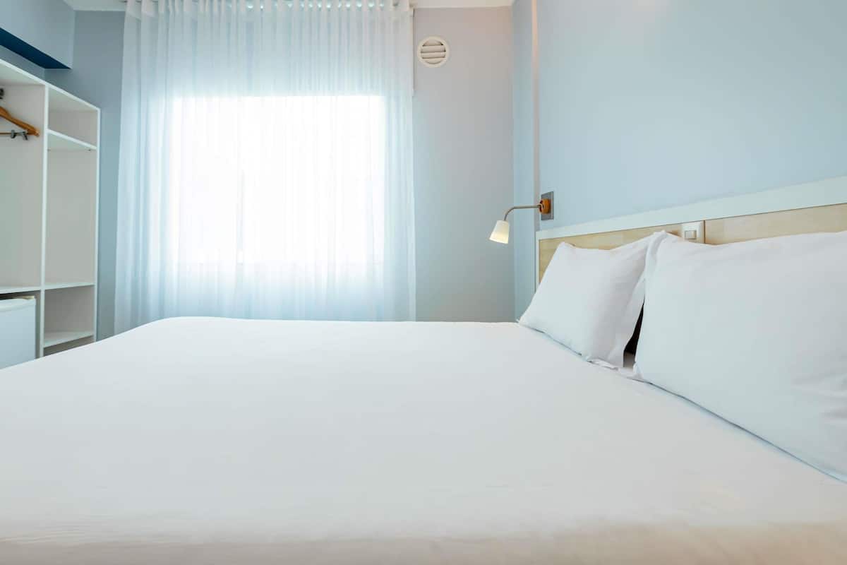 Una habitación de hotel con una cama grande, un armario, una ventana con cortinas translúcidas y una lámpara fijada a la pared.