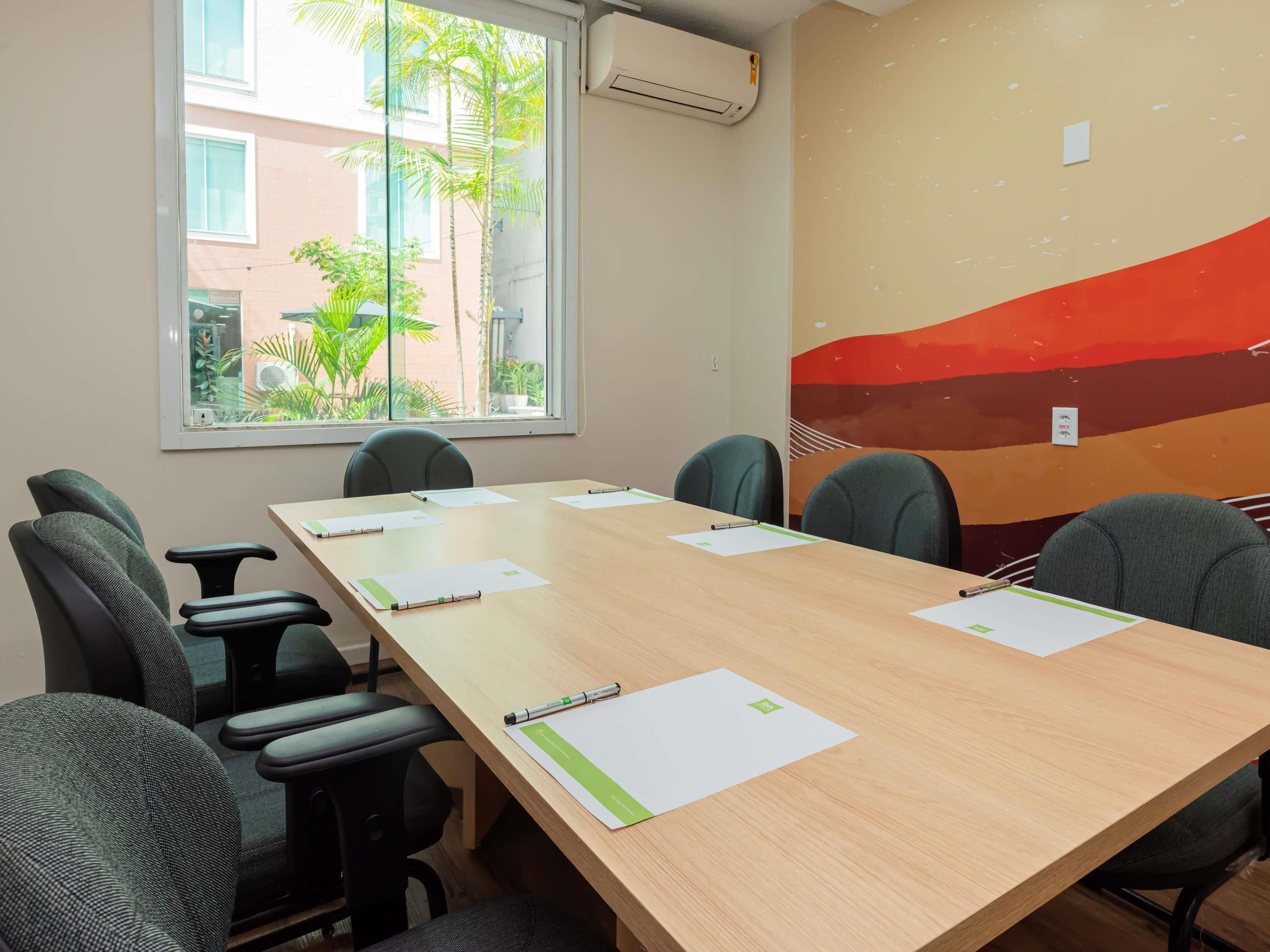 Sala de conferências com uma mesa comprida, seis cadeiras e uma janela com vista para edifícios e áreas verdes.