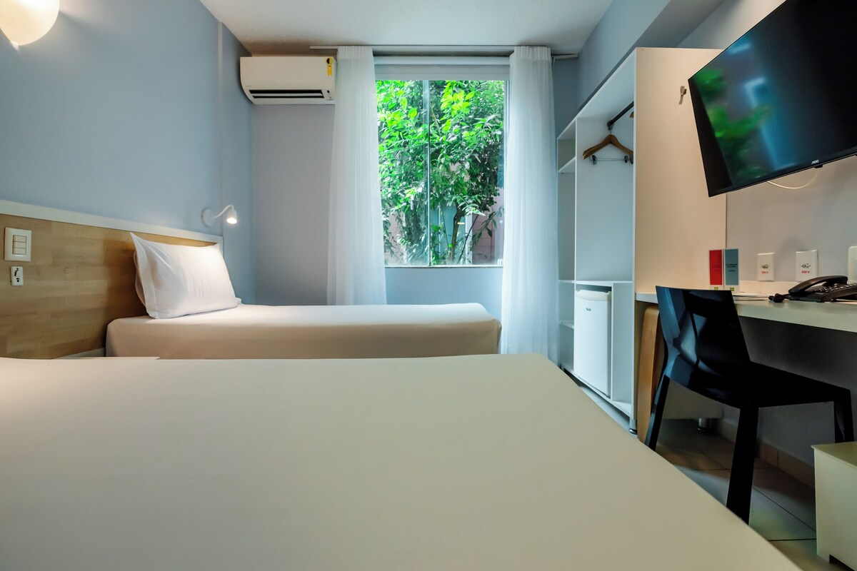 A hotel room with a bed, a desk with a chair, a television, and a window with a view of greenery.