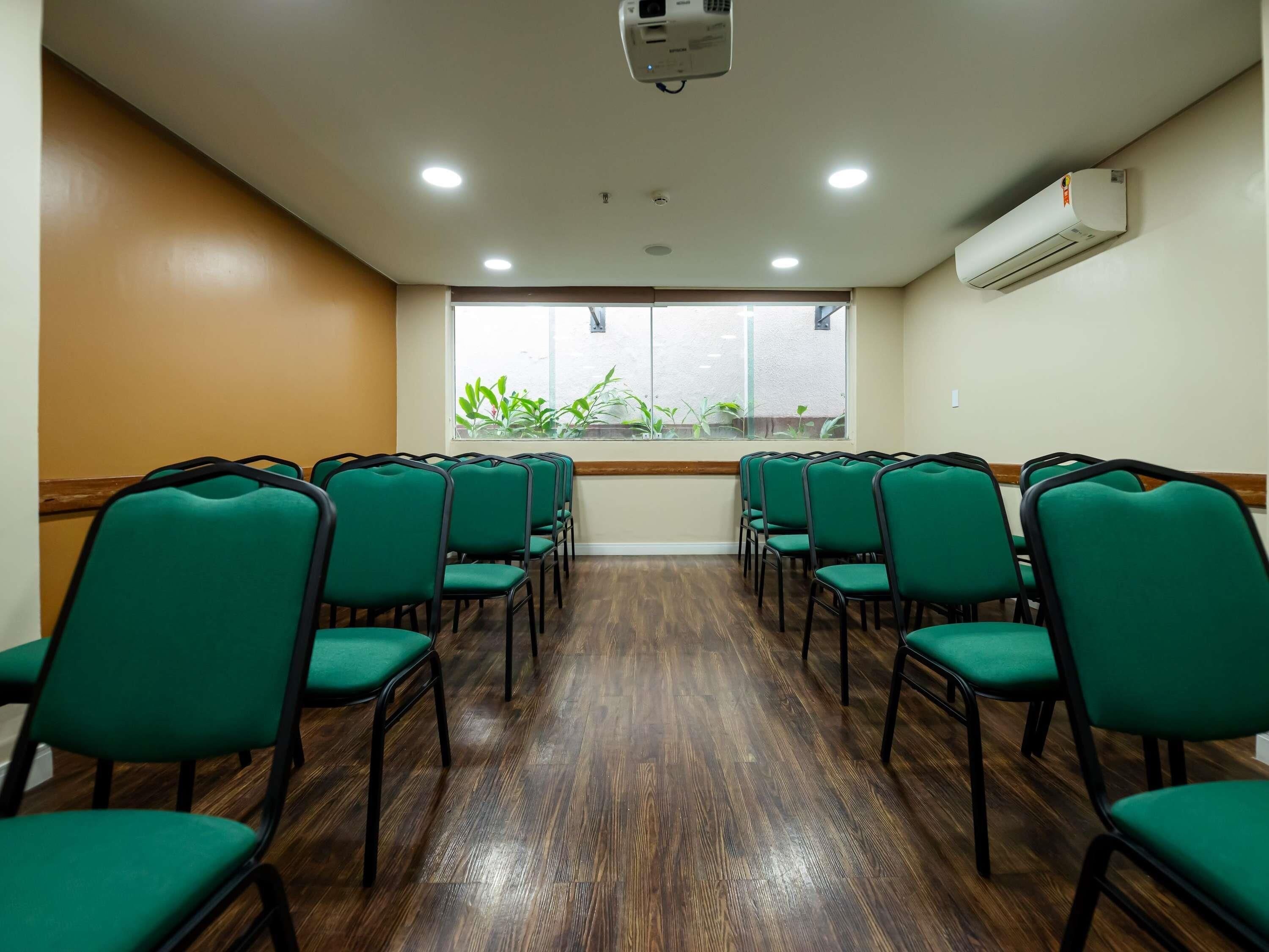 Un salón de conferencias con hileras de sillas verdes, suelo de madera, un ventanal con plantas y un equipo de aire acondicionado en la pared.