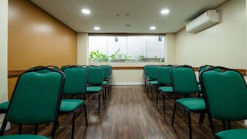 Un salón de conferencias con hileras de sillas verdes, suelo de madera, un ventanal con plantas y un equipo de aire acondicionado en la pared.