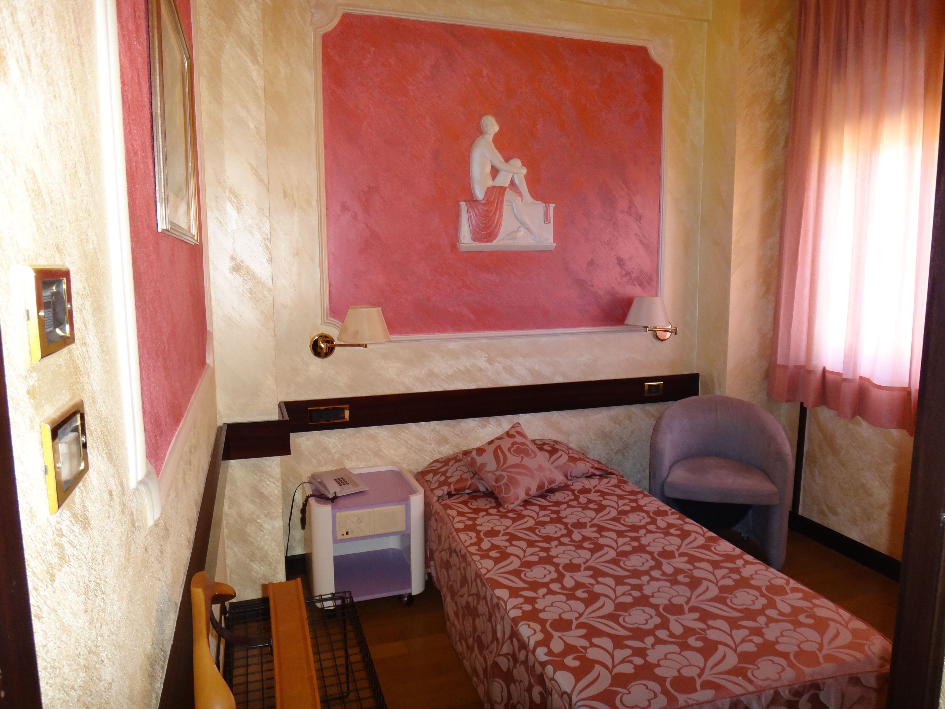 A small, compact room with a single bed, a chair, a bedside table, and a wall-mounted lamp.
