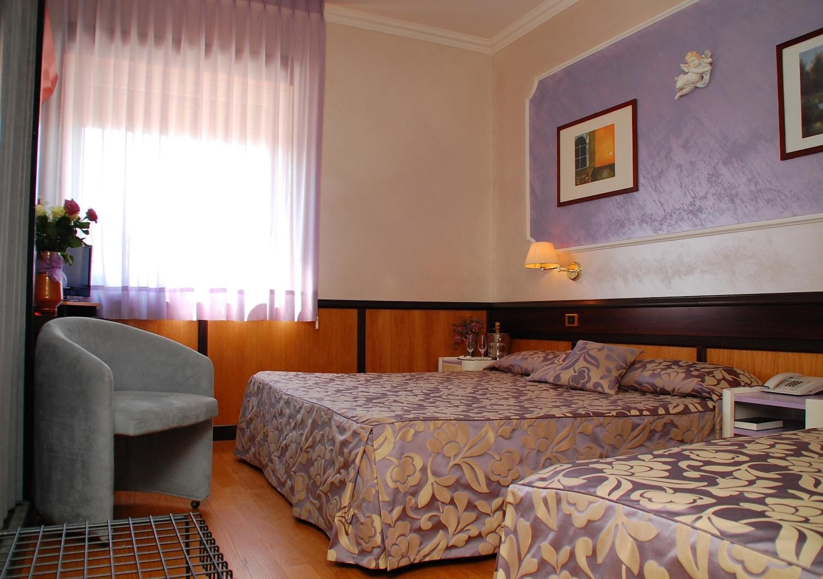 Habitación de hotel con dos camas, una silla, un ventanal con cortinas, una mesita de noche y una lámpara fijada a la pared.
