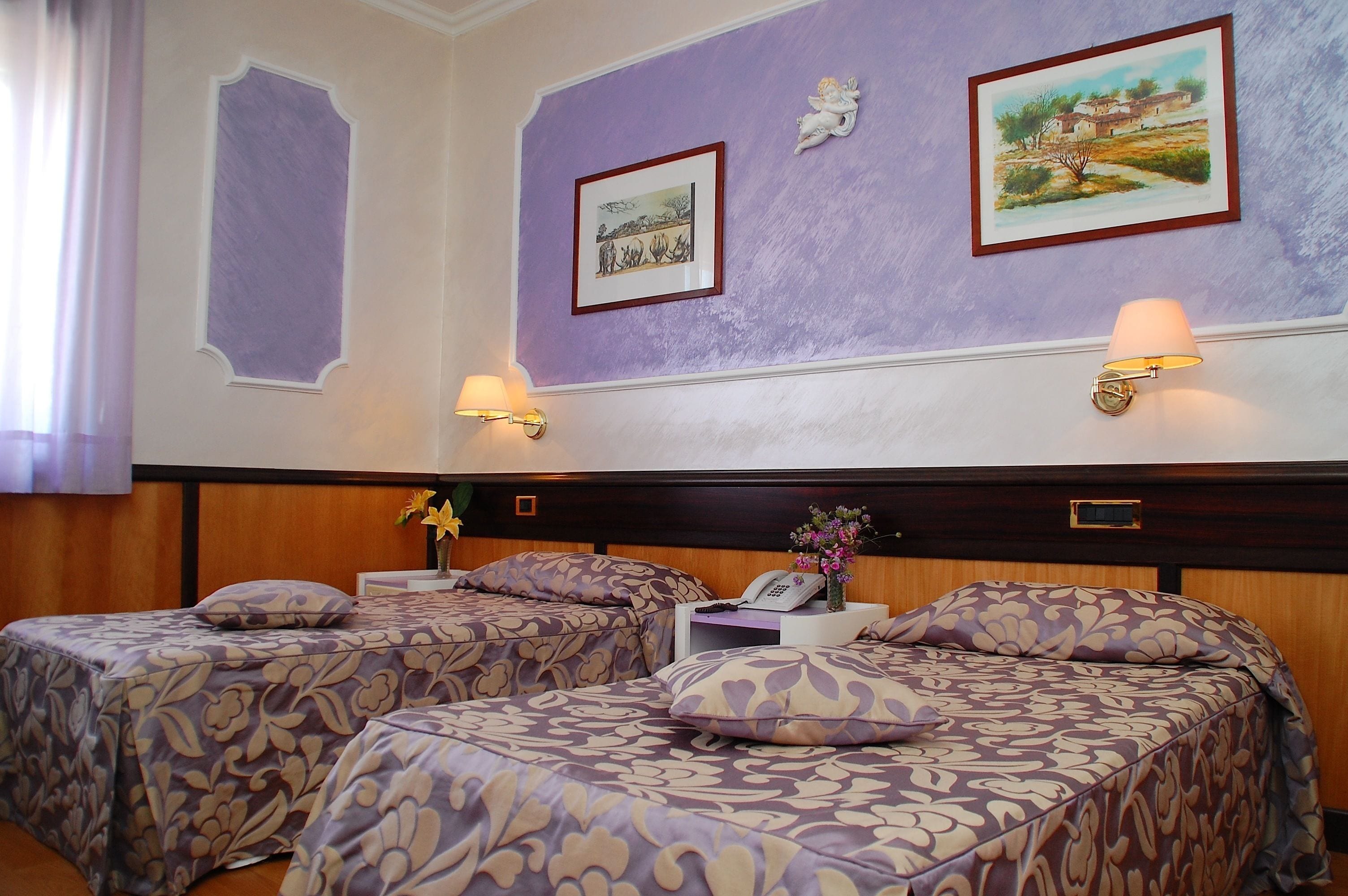 A hotel room with two beds, each with patterned bedding, a nightstand with a lamp, and artwork on the walls.