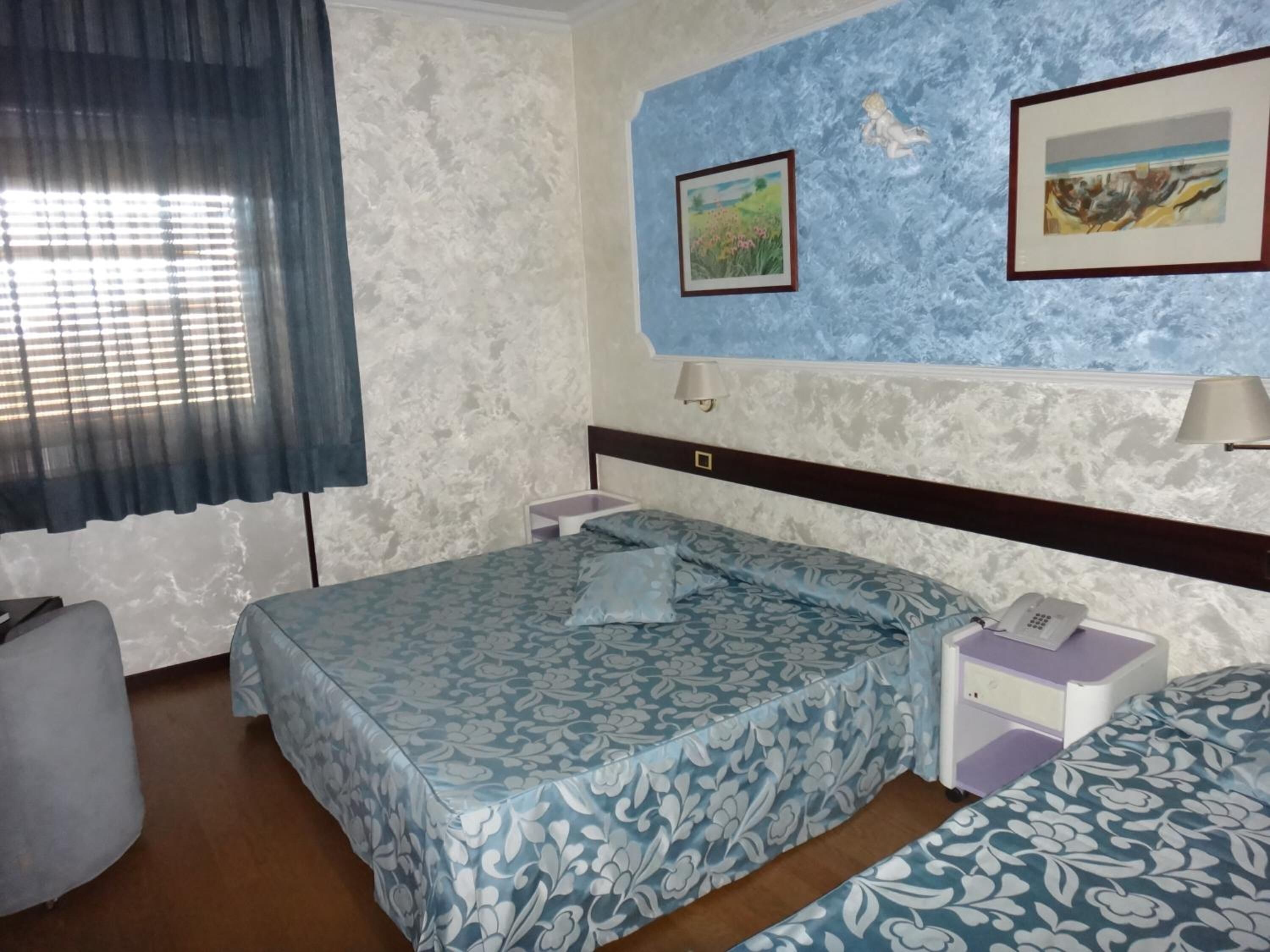 Ein Hotelzimmer mit einem Bett, einem Nachttisch mit Lampe, einem kleinen Tisch mit Telefon und einem Fenster mit Vorhängen.