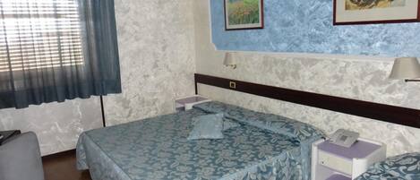 A hotel room with a bed, a bedside table with a lamp, a small table with a phone, and a window with curtains.