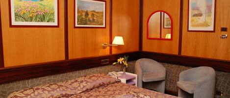 Habitación de hotel con cama, dos sillones, mesita de noche con lámpara y cuadros en las paredes.