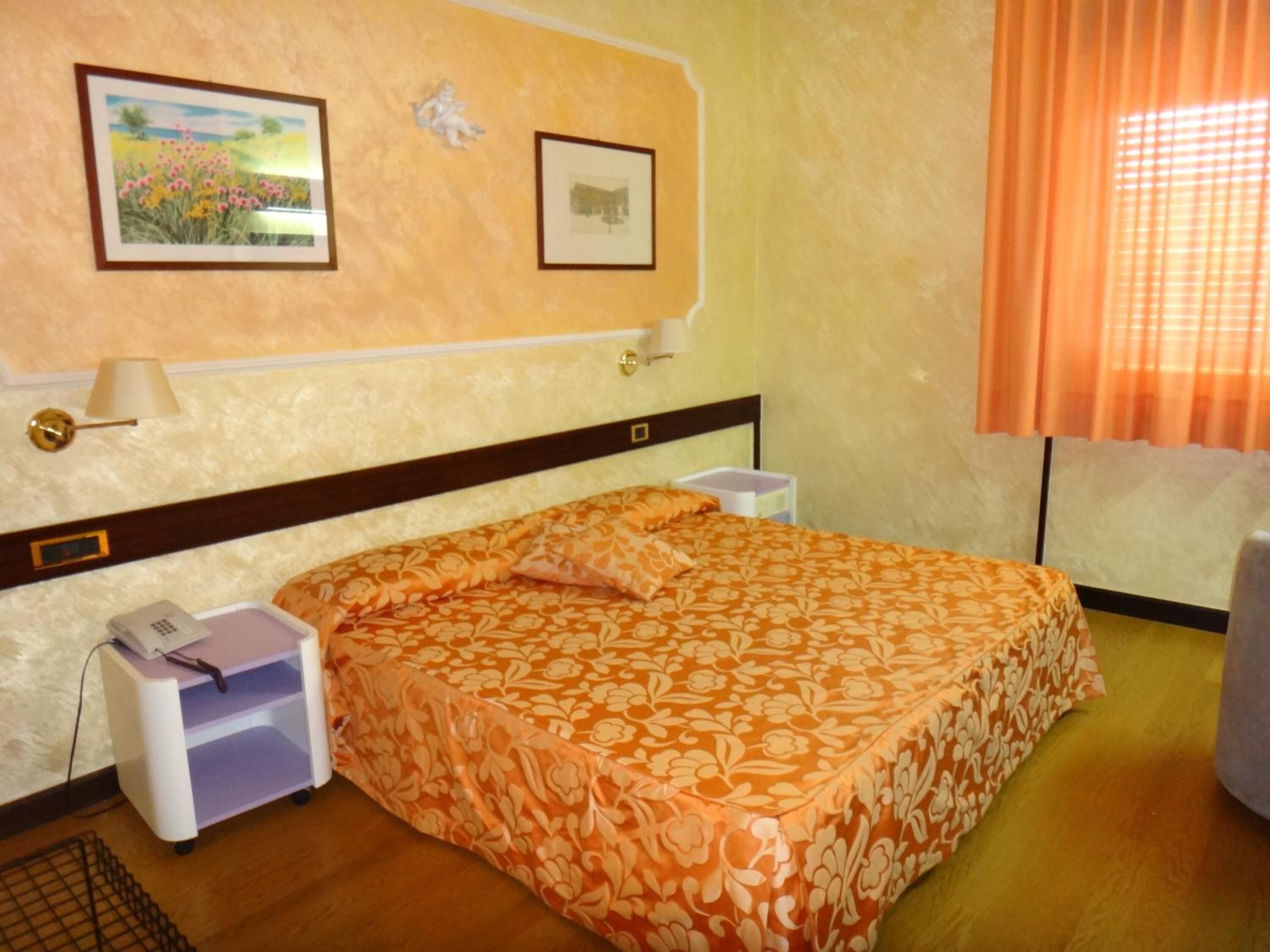 A bedroom with a floral bedspread, a nightstand with a phone, a wall-mounted lamp, and two framed pictures.