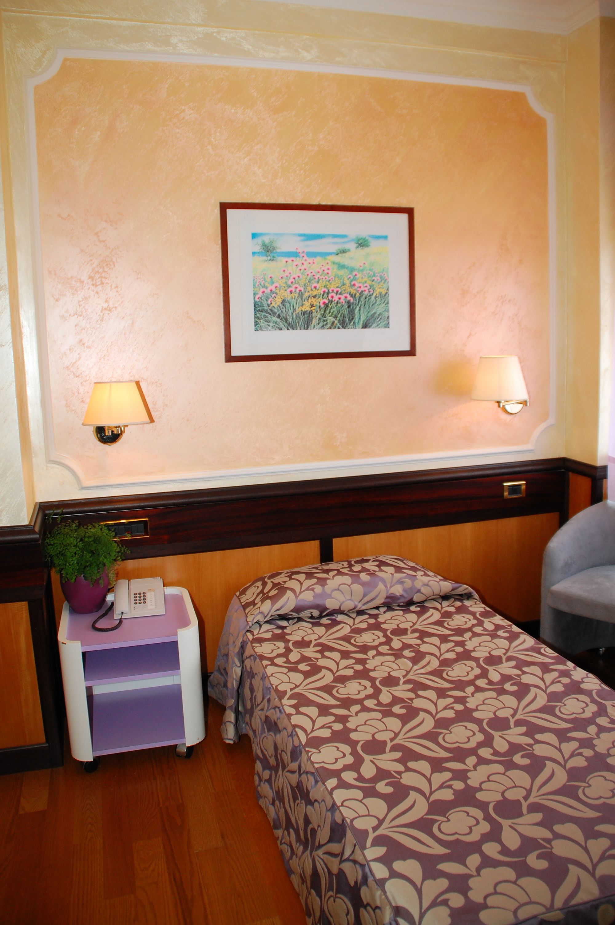 A hotel room with a bed, bedside lamps, a small table with a plant, a telephone, and a framed picture on the wall.