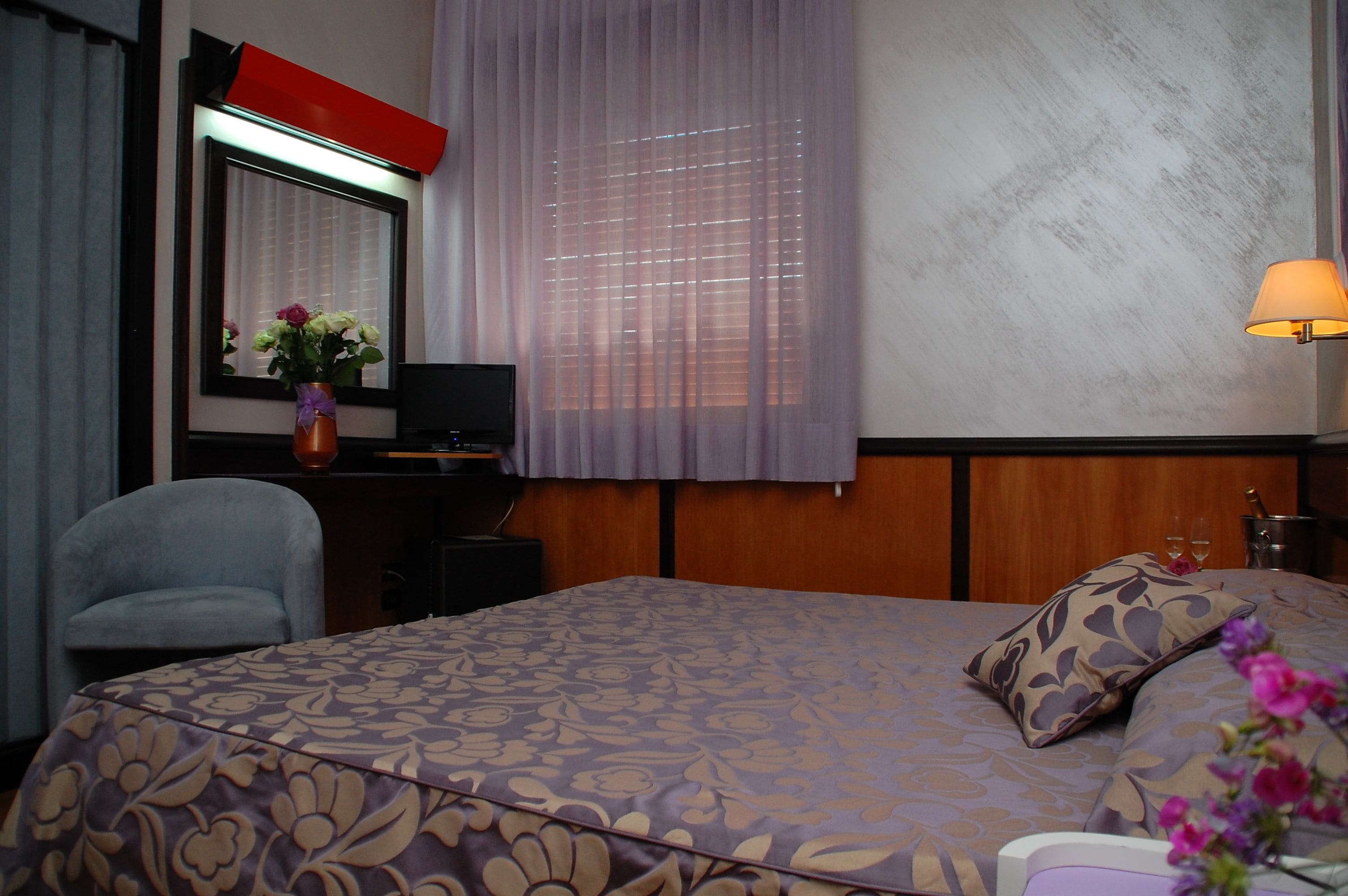 Habitación de hotel con cama, escritorio, silla, televisor, lámpara y ventana con cortinas.