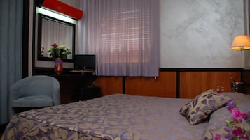 Habitación de hotel con cama, escritorio, silla, televisor, lámpara y ventana con cortinas.