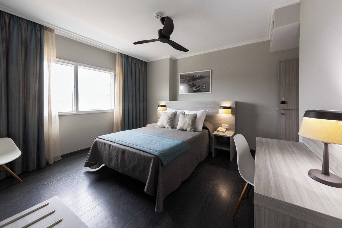 A modern hotel room with a bed, bedside tables, a desk, and a ceiling fan.