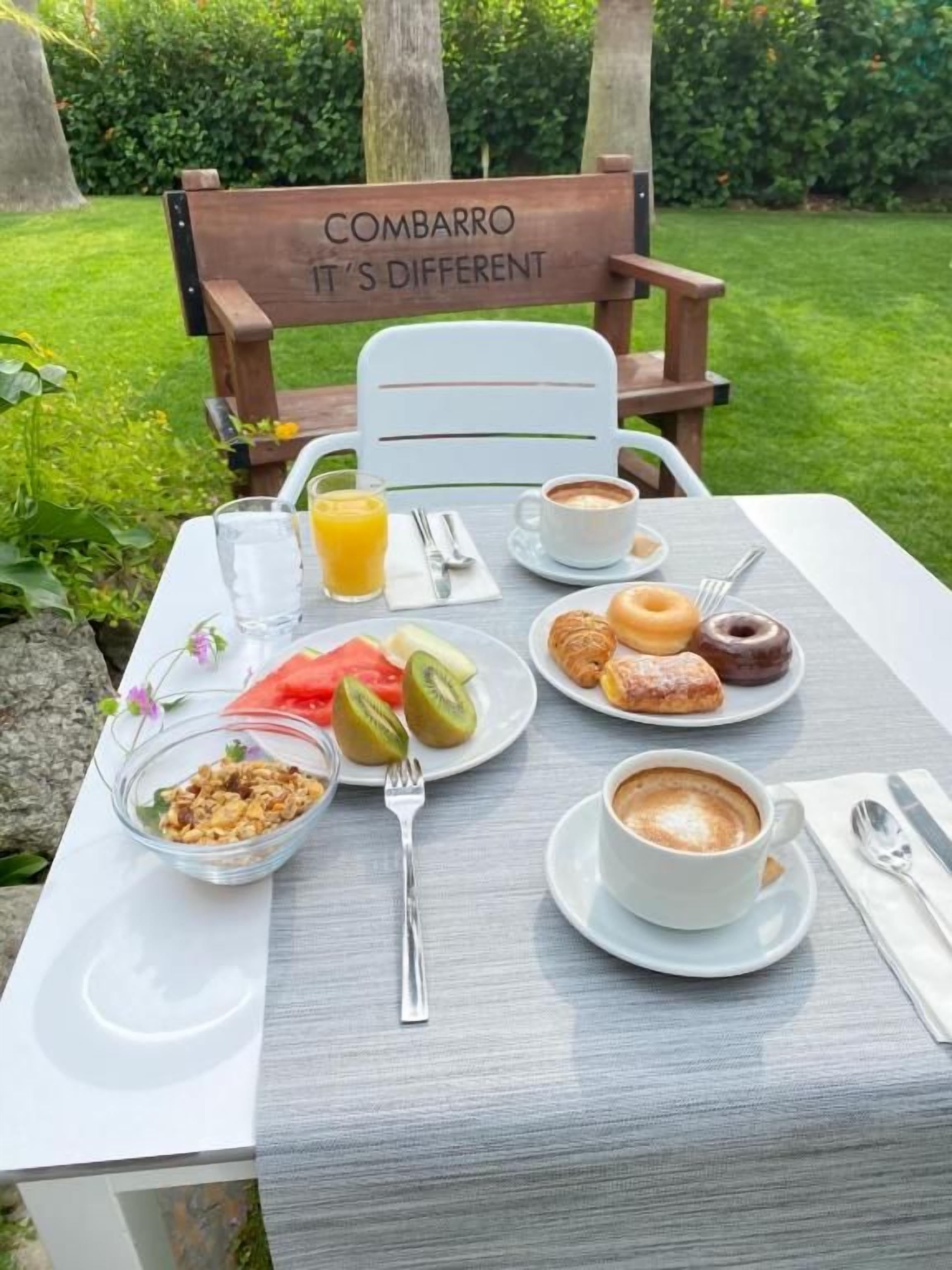 A table set outdoors with a sign reading 'COMBARRO IT'S DIFFERENT', a breakfast spread including toast, fruit, and coffee, and a chair with the same sign.