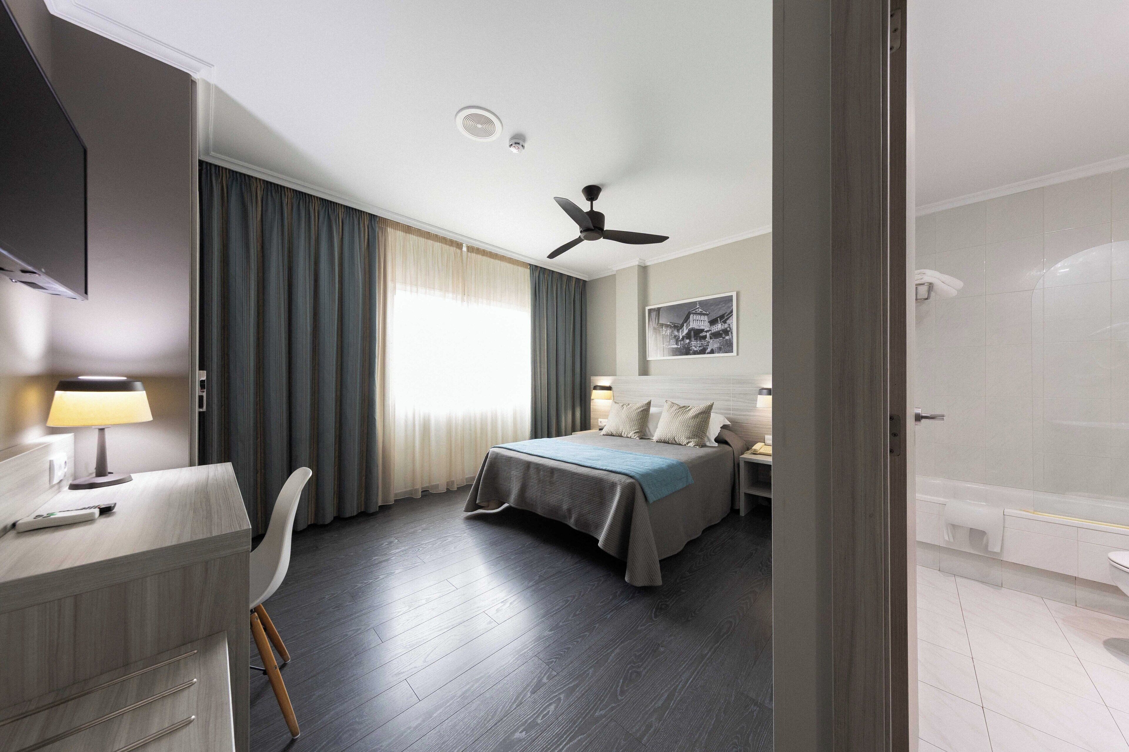 A hotel room with a bed, a desk, a chair, a ceiling fan, and a bathroom with a bathtub and toilet.