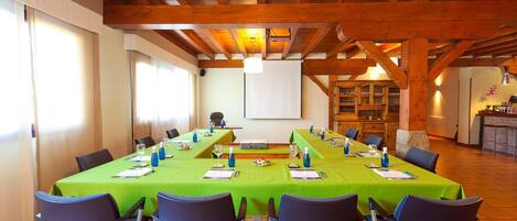 Un salón de conferencias con una mesa larga, sillas, una pantalla de proyector y una cocina al fondo.