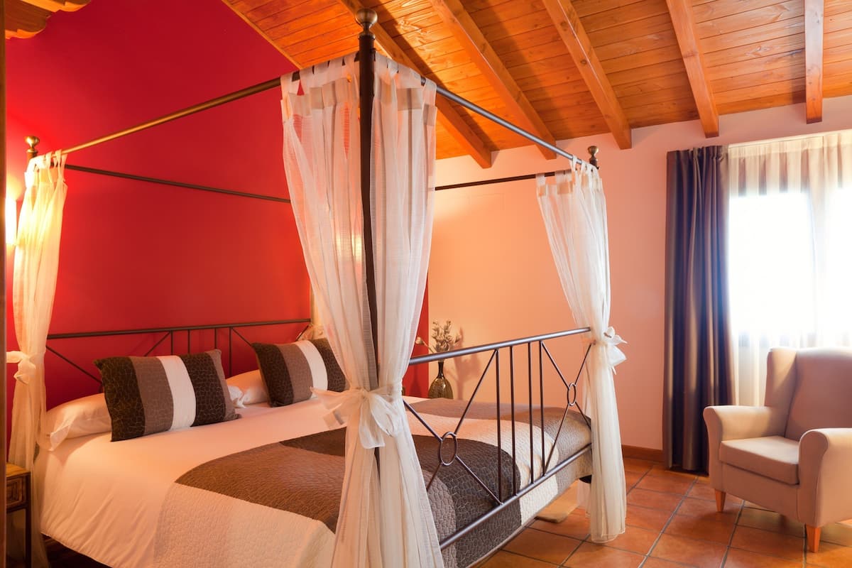 A four-poster bed with white curtains, a wooden ceiling, and a red wall.