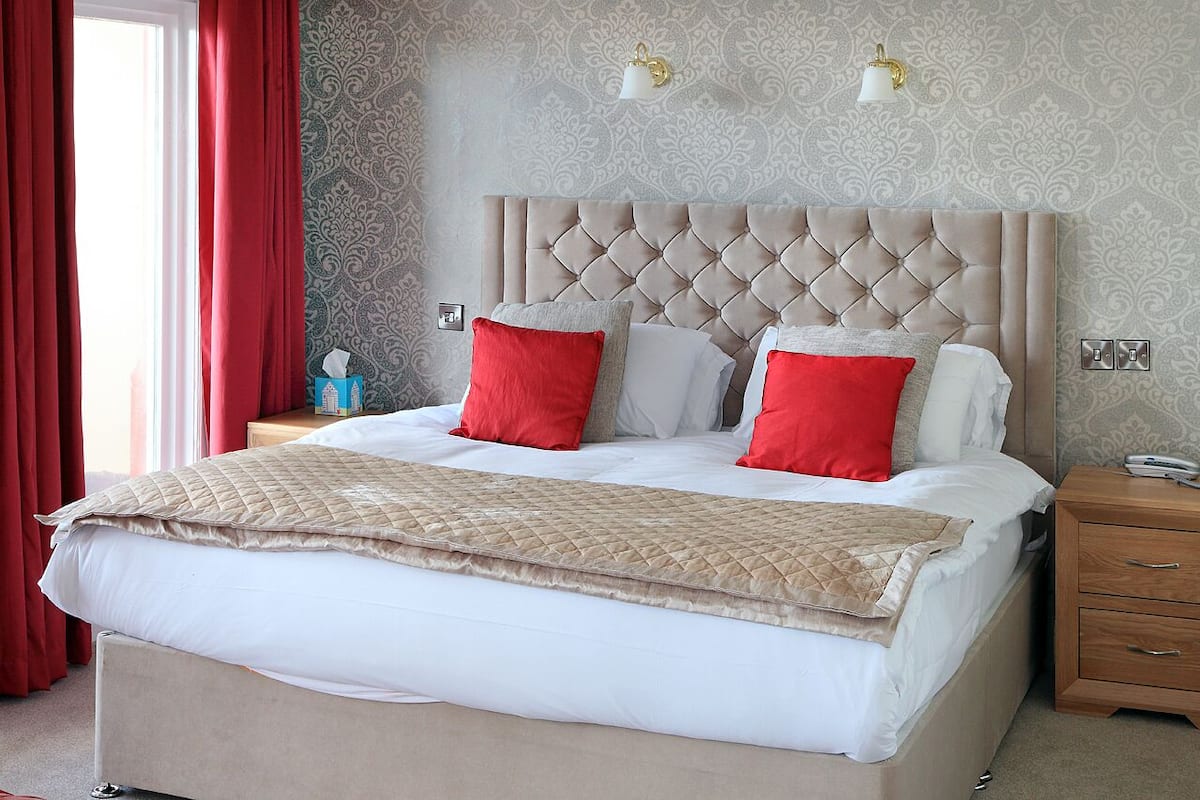 Uma cama bem arrumada com um edredom acolchoado e duas almofadas vermelhas, uma mesinha de cabeceira de madeira com um abajur e um papel de parede estampado.