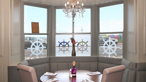 Un espace repas avec une table ronde, des banquettes grises rembourrées, un lustre et de grandes fenêtres décorées de motifs de roue de navire.
